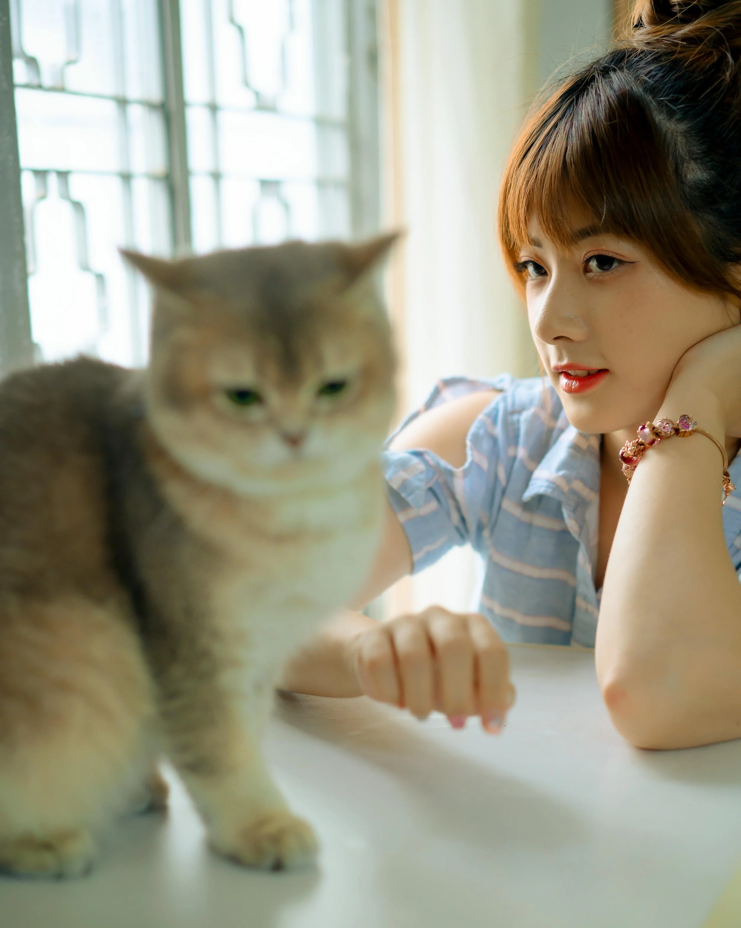 A young woman with short hair and a striped shirt resting her chin on her hand, looking at a cat on a table by a window.