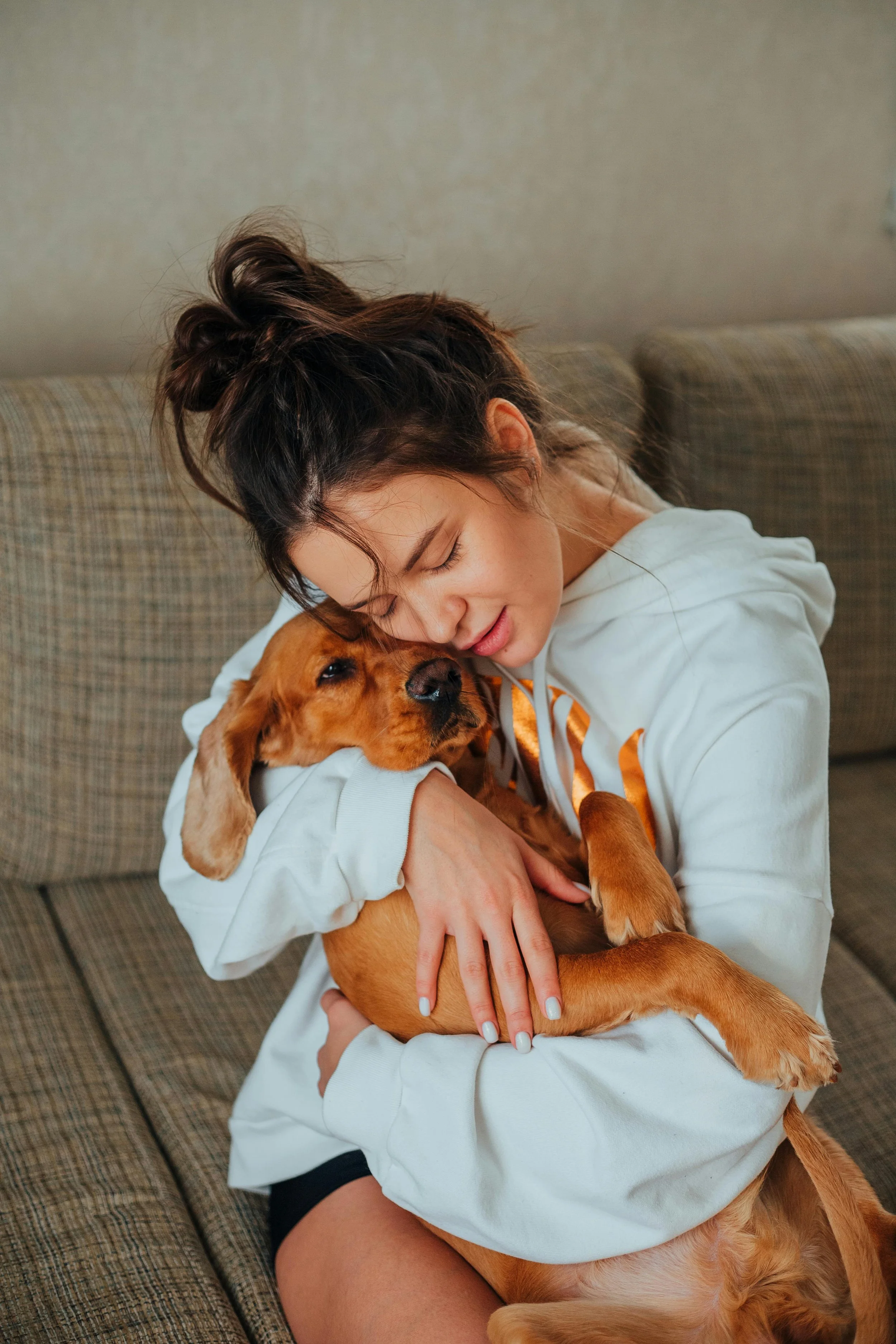 A woman with dark hair in a bun hugging a brown puppy on a brown checkered couch.