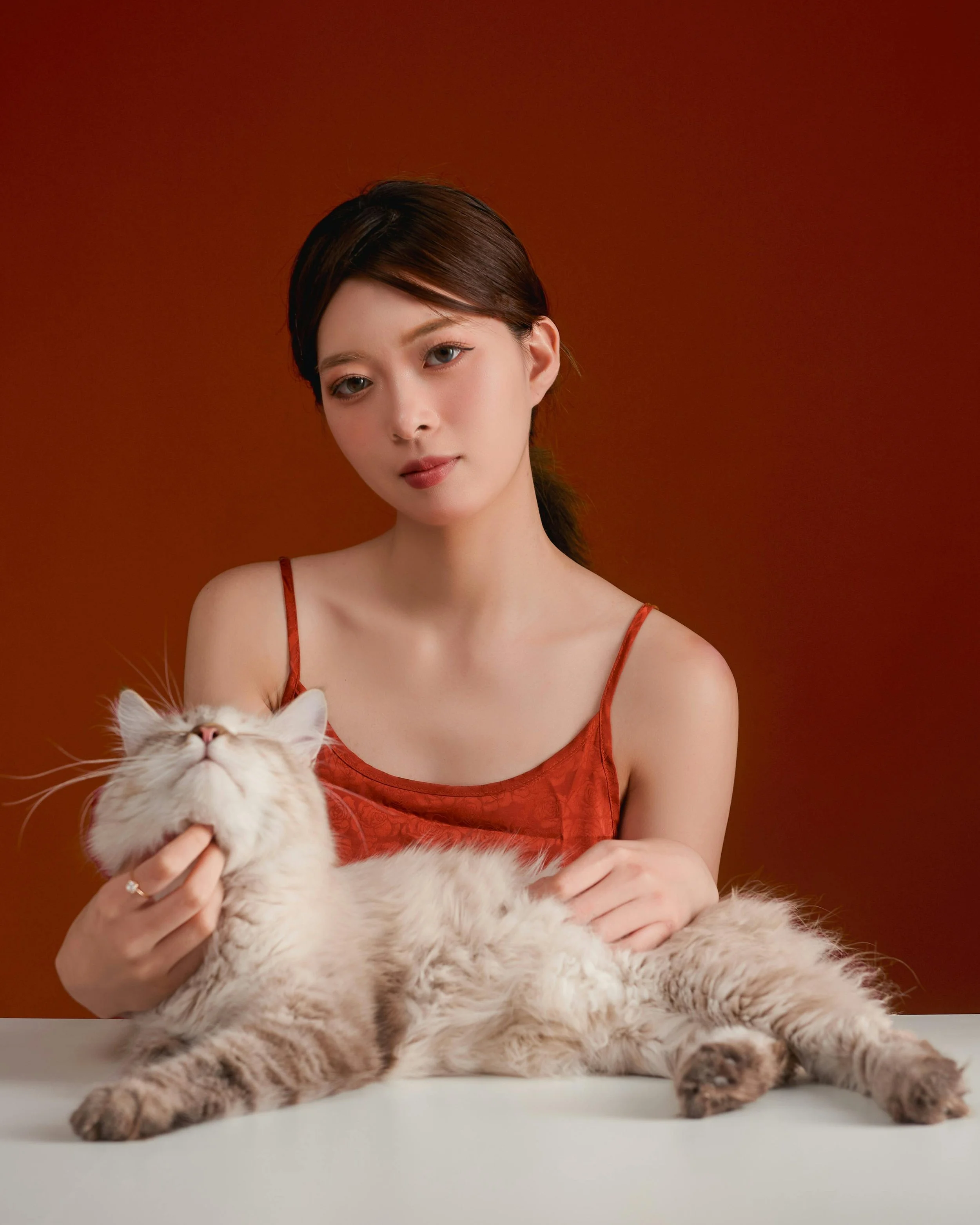 A woman with brown hair and fair skin holding a fluffy cream-colored cat with closed eyes, against a warm reddish-brown background.