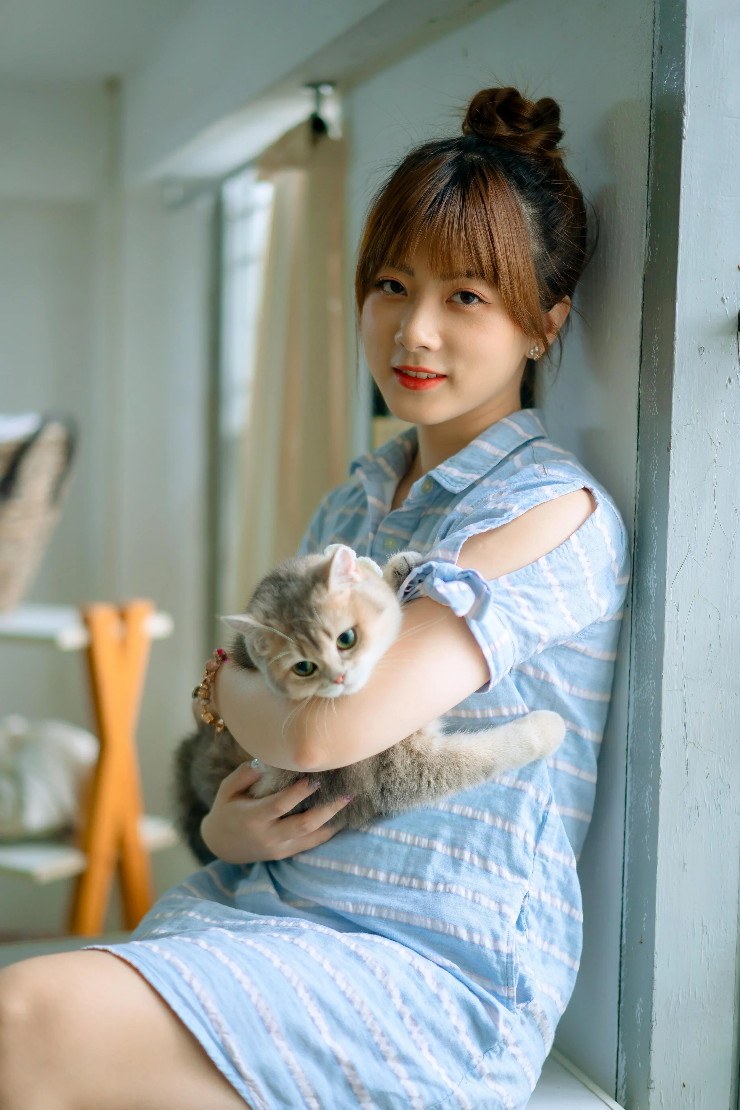 A young woman with brown hair in a bun and wearing a light blue stripped dress is sitting against a wall, holding a gray tabby cat with green eyes. She is smiling at the camera in a bright, cozy interior.
