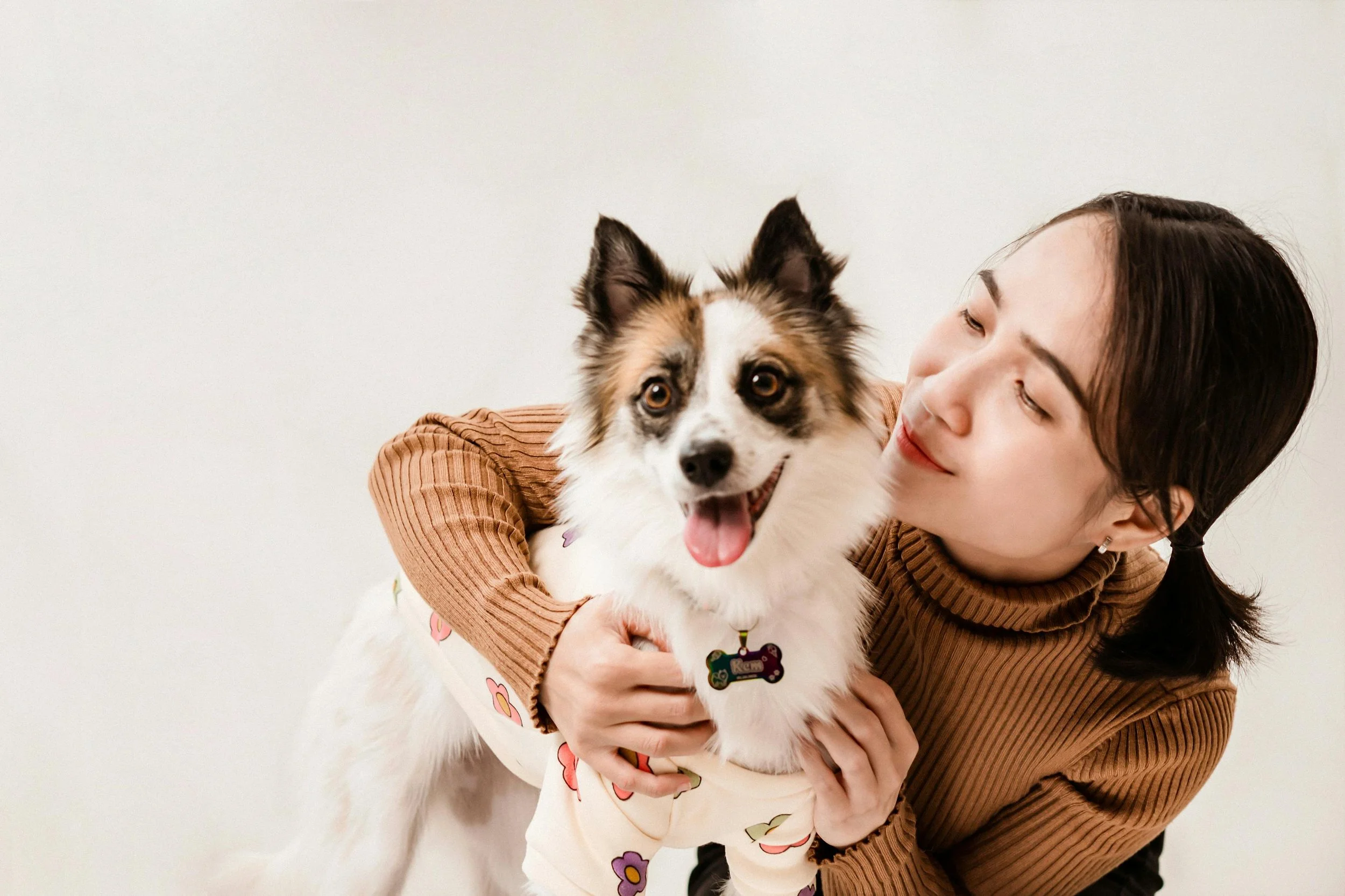 Young woman with dark hair and a brown ribbed turtleneck hugging a happy Australian Shepherd dog with a colorful collar and bone-shaped tag, against a plain white background.