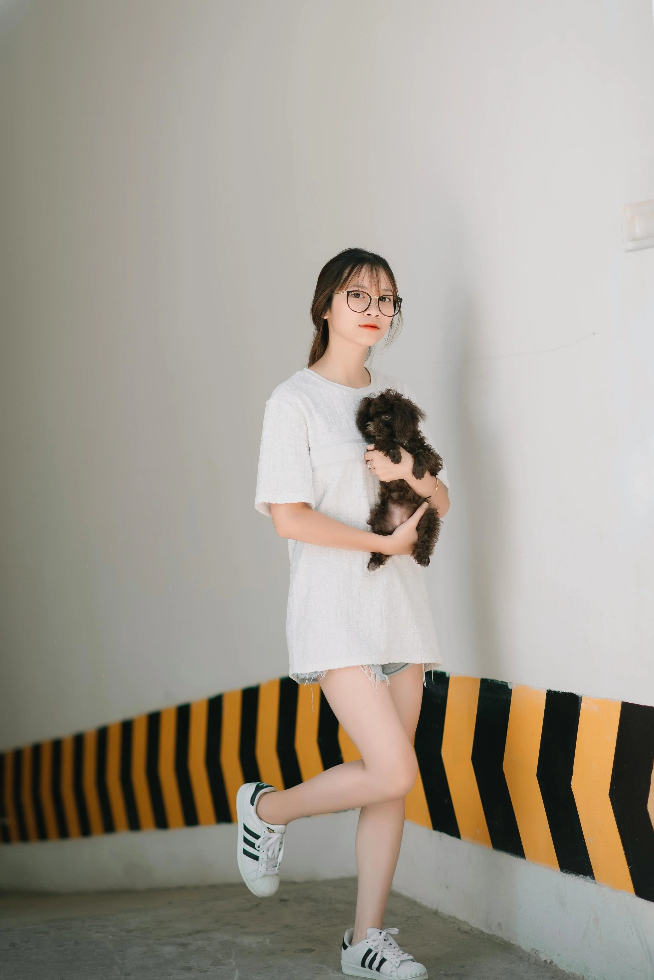 A young woman with long dark hair, glasses, and light skin, holding a small black puppy, standing in an indoor parking garage or similar space with a white wall and a black-and-yellow striped safety barrier.