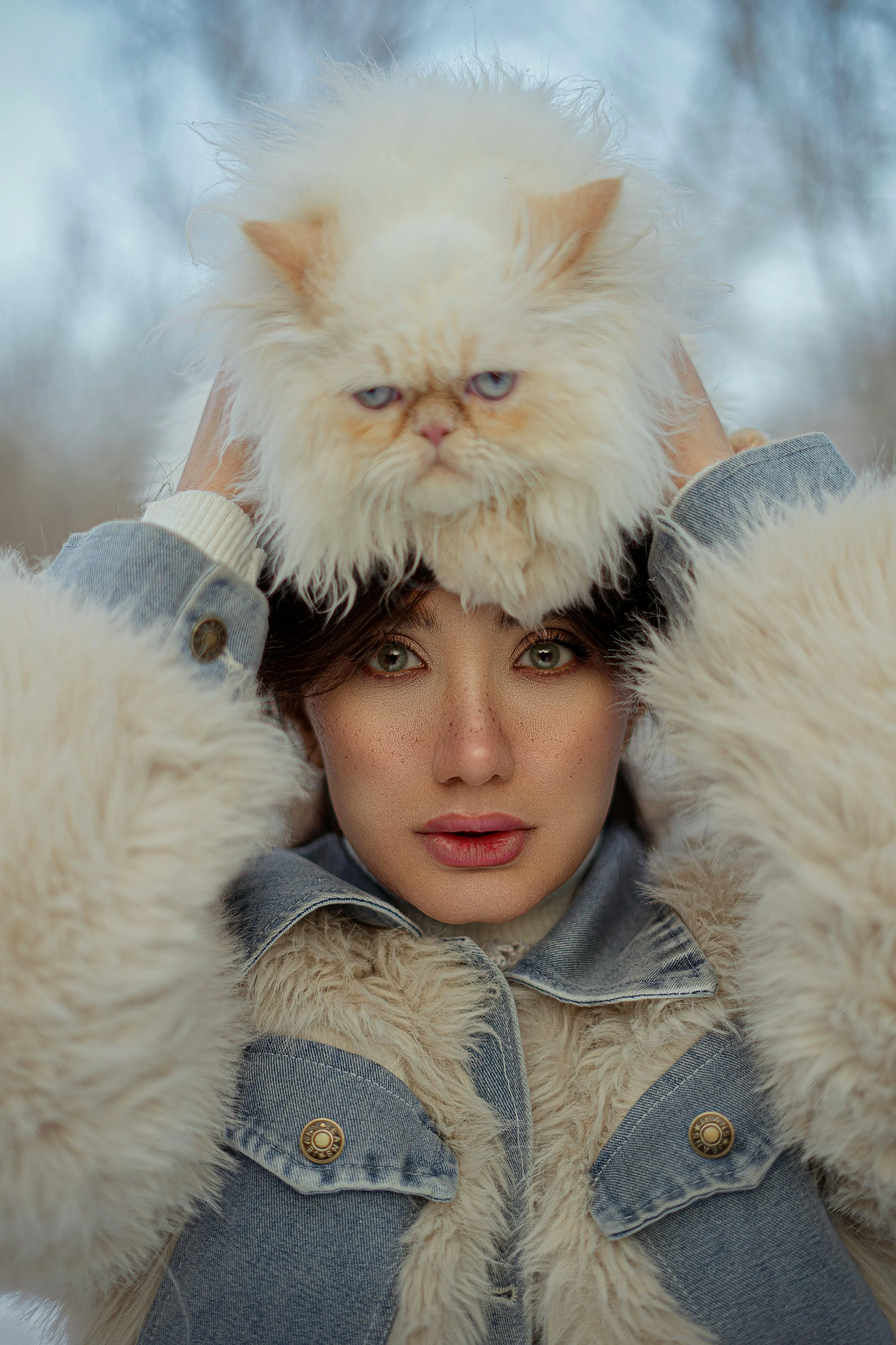 A woman with green eyes and freckles holding a large, fluffy, cream-colored cat on her head outdoors during winter.