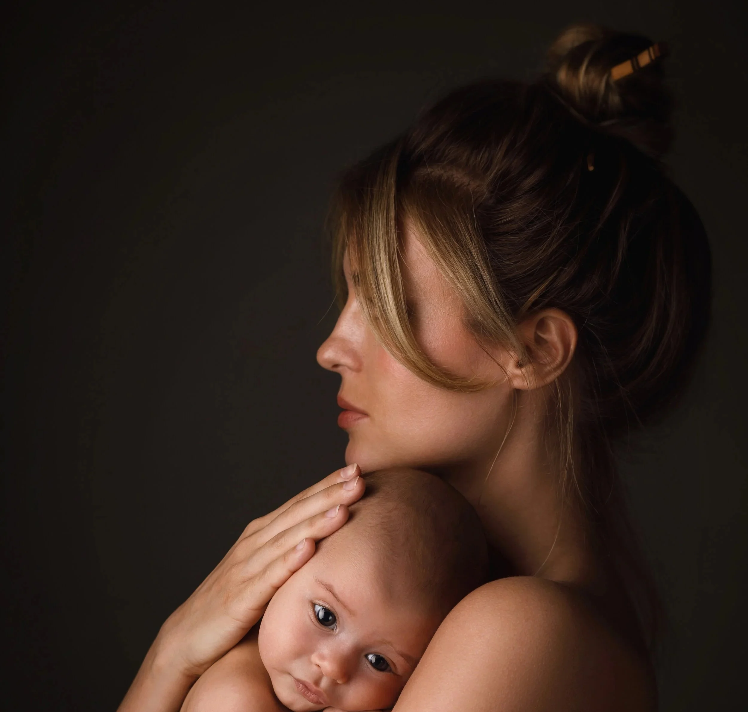 A woman gently holding a baby with light skin against her shoulder, both with relaxed expressions, against a dark background.