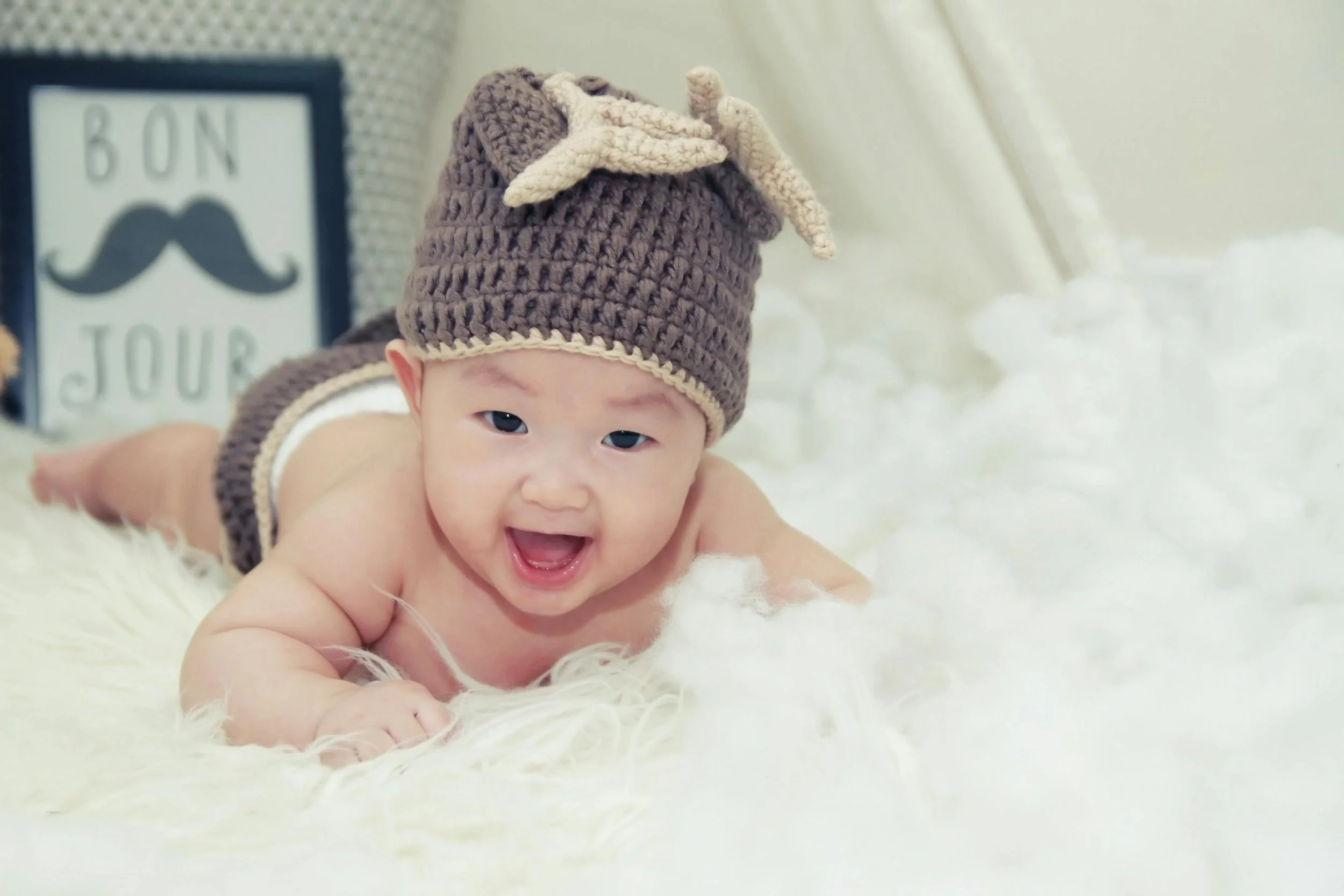 A smiling baby crawling on a soft, white fuzzy surface, wearing a brown handmade crochet elf hat with a beige bow, with a picture of a mustache and the words 'Bon Jour' in the background.