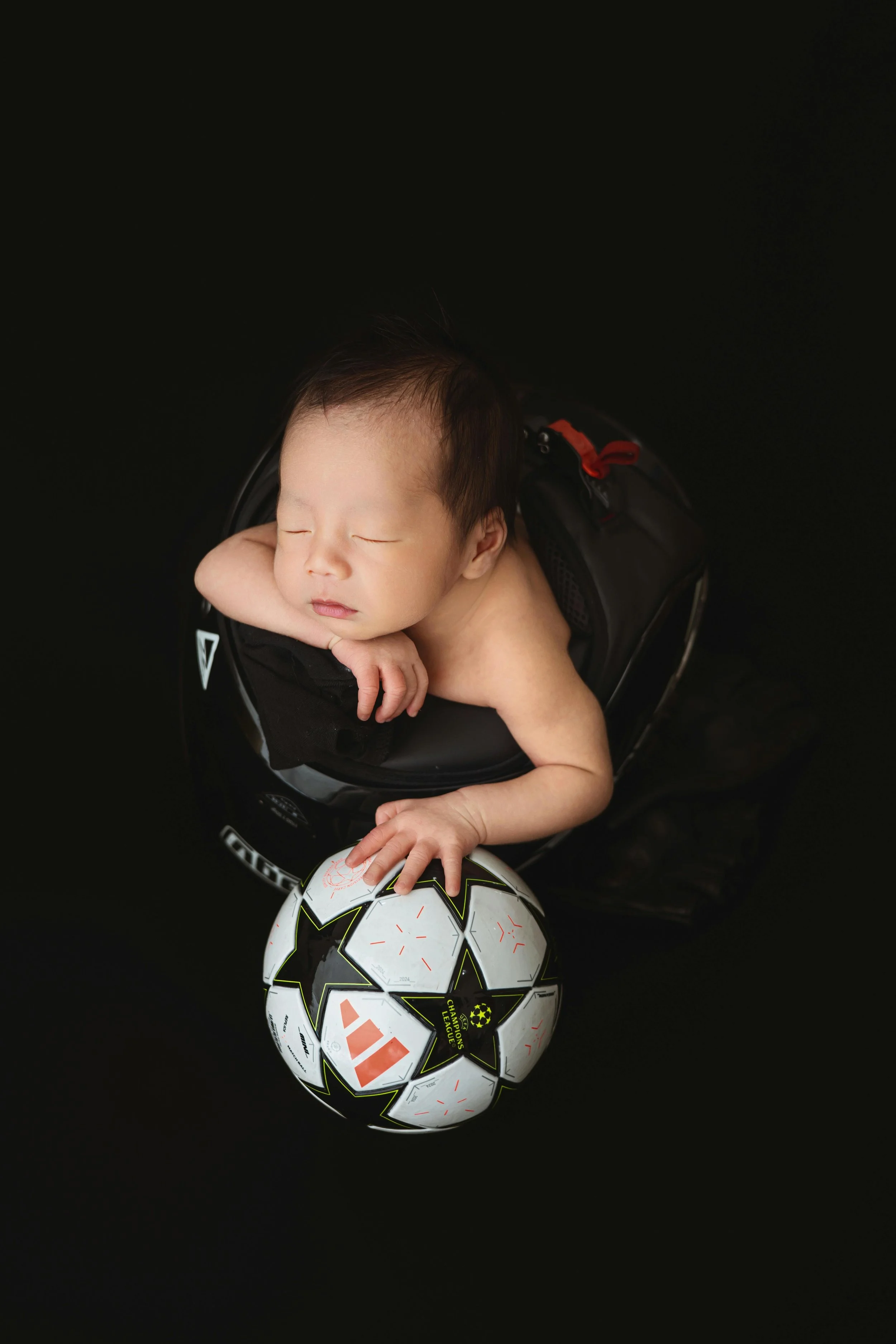 Young baby sleeping on a black car seat with a soccer ball nearby.