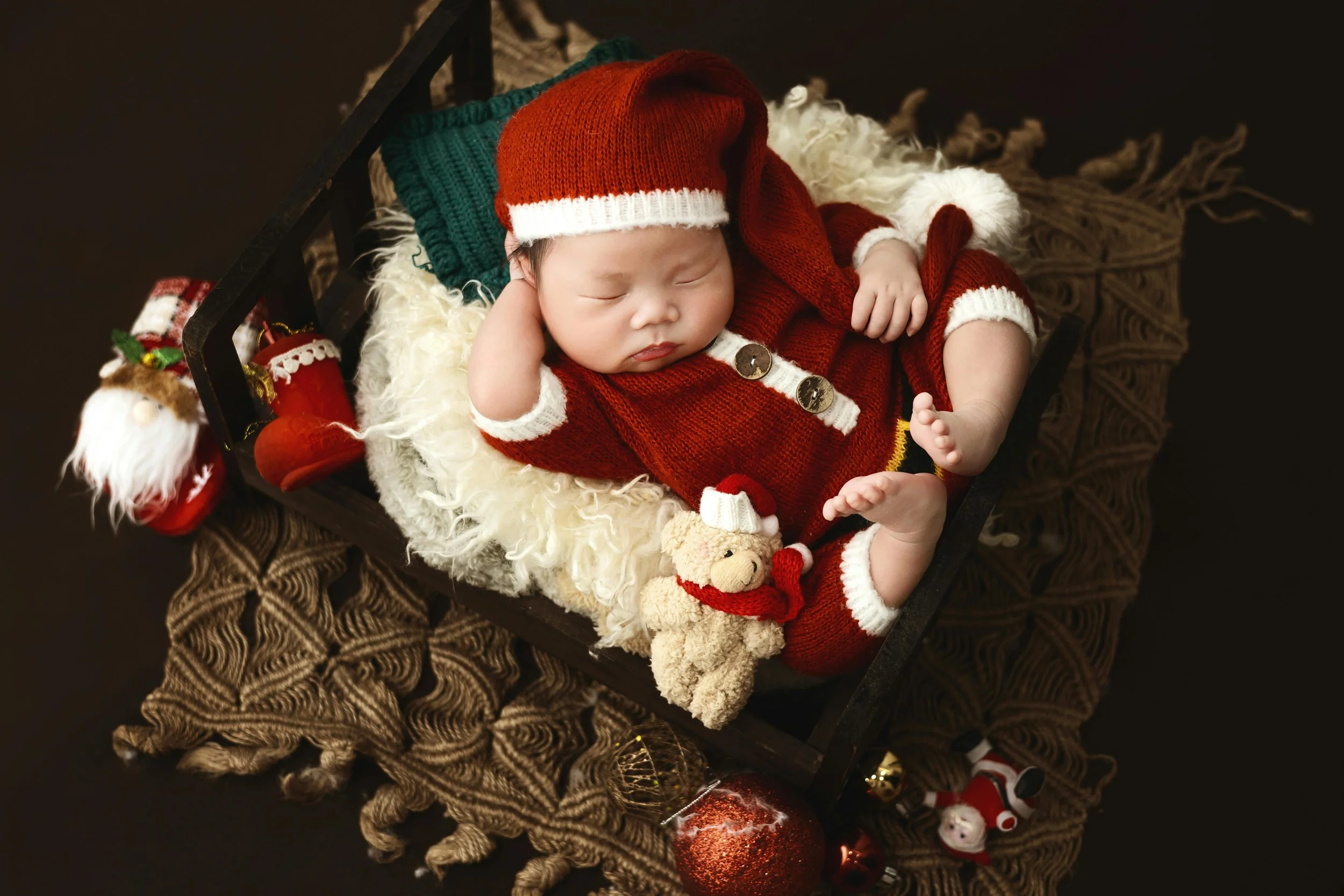 A sleeping baby dressed in a red Christmas outfit with white trim and buttons, wearing a matching red and white Santa hat, lying in a small wooden bed surrounded by Christmas decorations and plush toys, with a dark background.