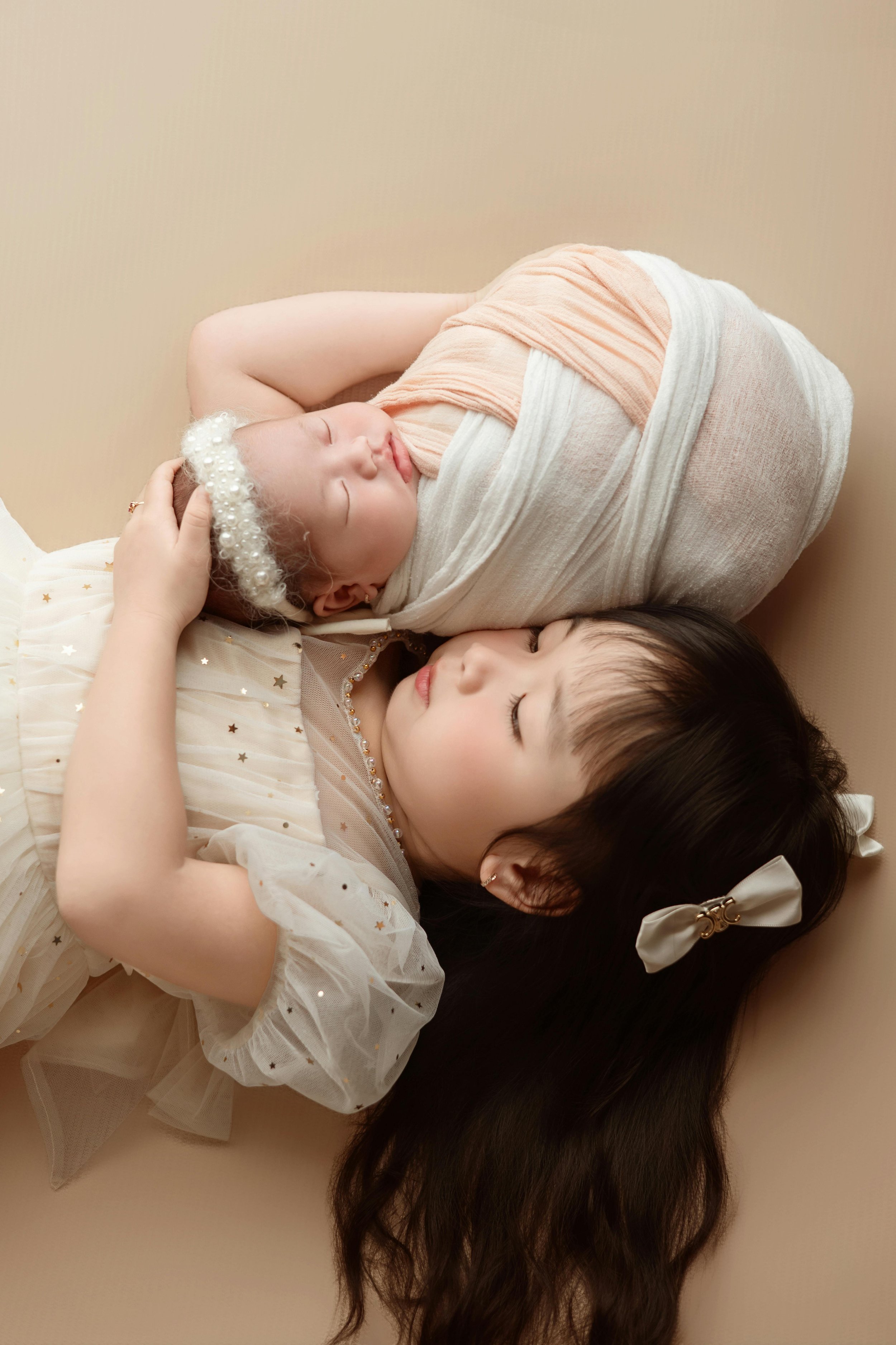A young girl lying down with eyes closed, gently holding the head of a sleeping baby wrapped in a blanket. The girl has long dark hair with a bow and wears a white dress with sheer sleeves decorated with small sparkles. The baby has a pearl headband 