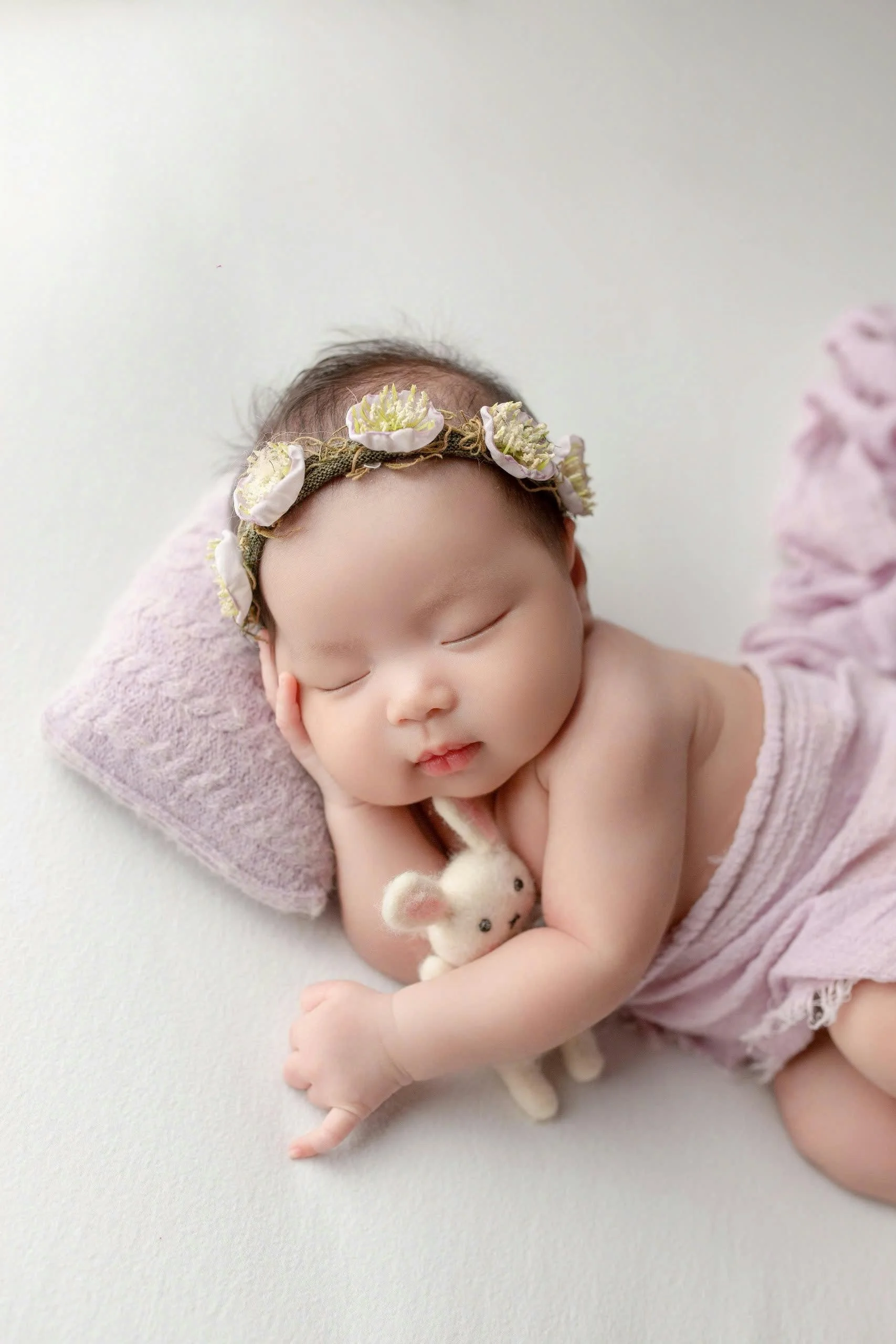 A sleeping baby girl with a floral headband, lying on a pink pillow with a tiny stuffed animal, wrapped in a soft pink blanket.
