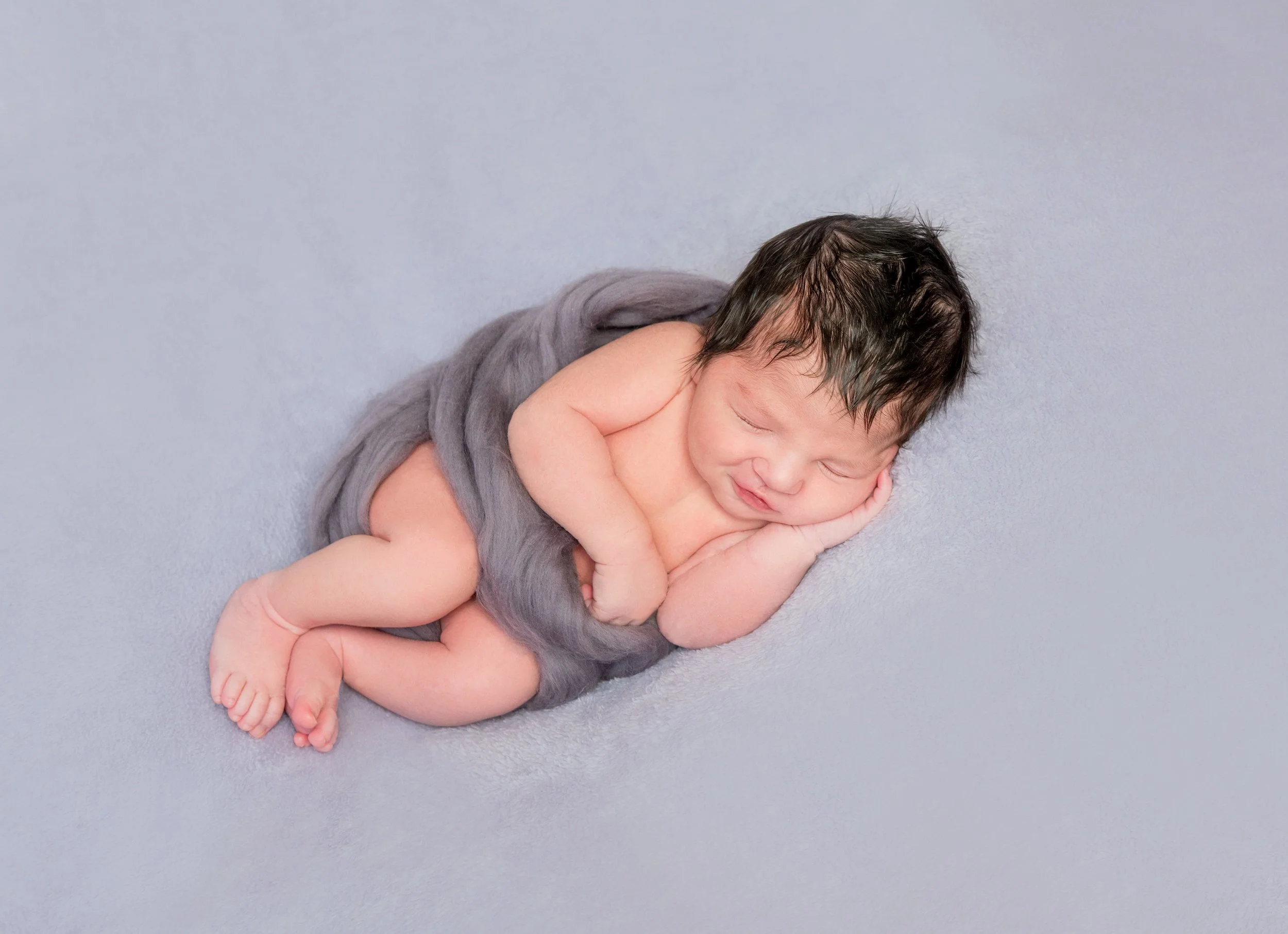 A sleeping newborn baby with dark hair, lying on a soft, light gray surface, wrapped partially in a gray cloth, with hands tucked under the head and a peaceful expression.