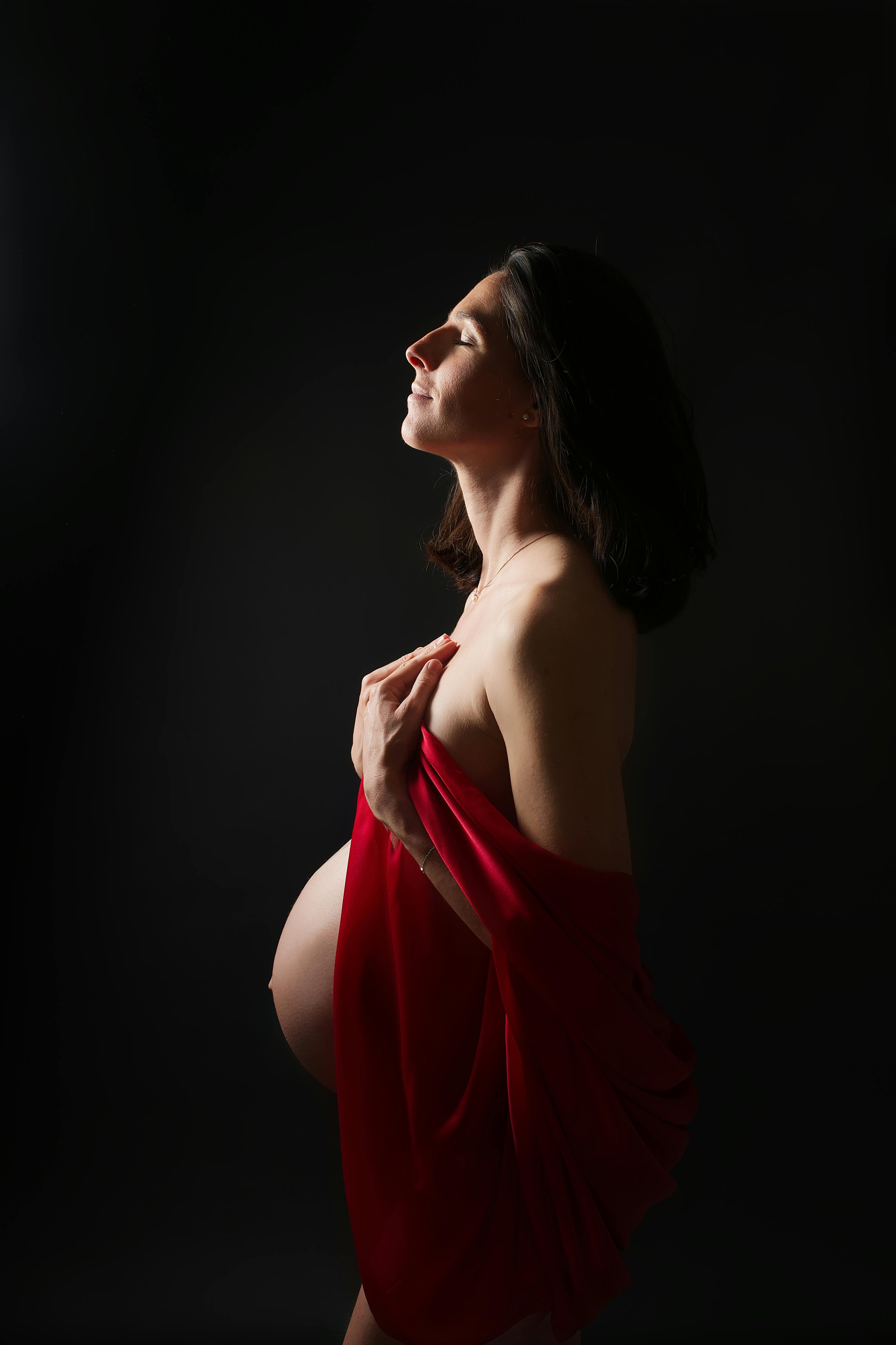 Pregnant woman with eyes closed, holding red fabric across her chest against a black background.