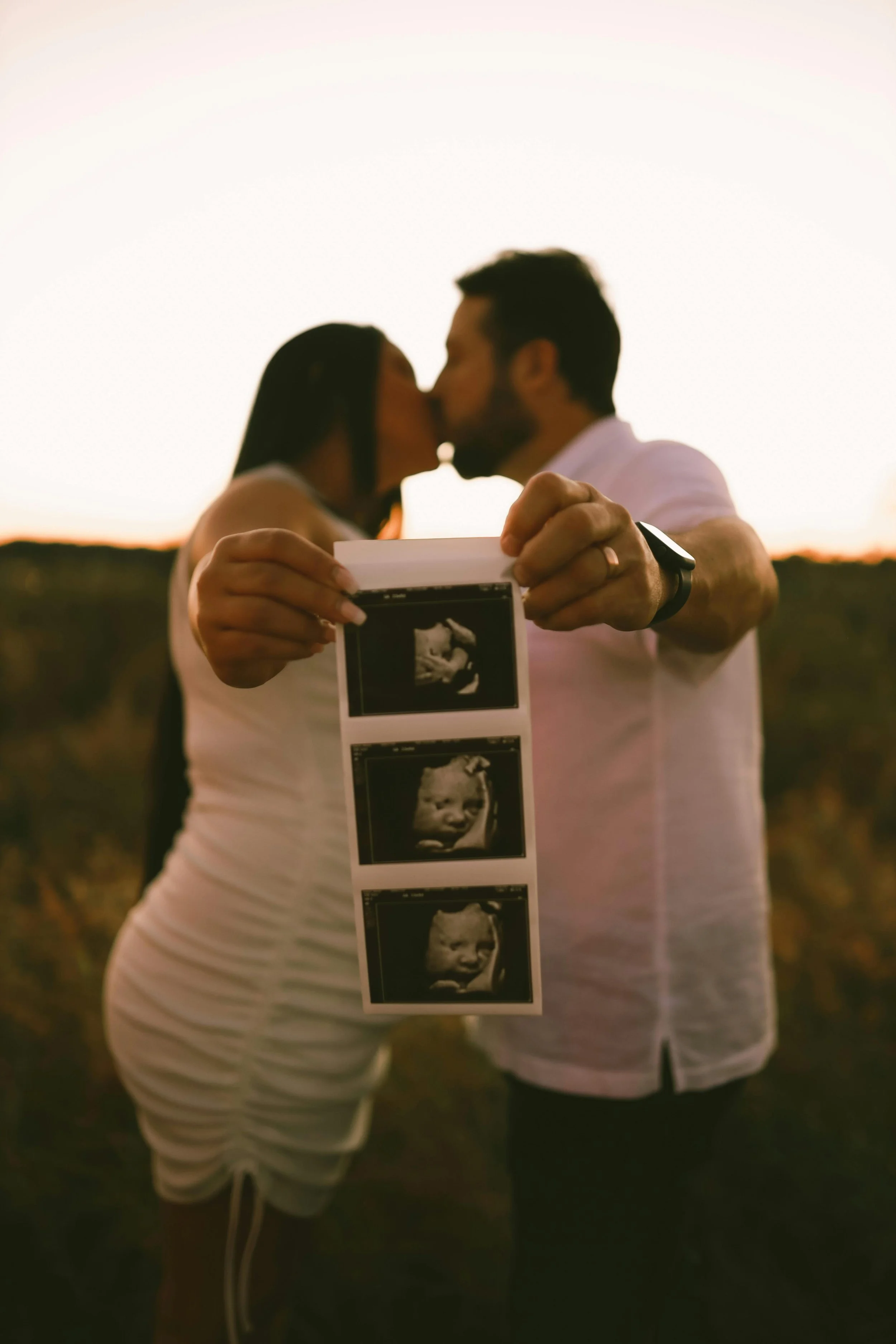 Couple holding ultrasound images and sharing a kiss outdoors during sunset.
