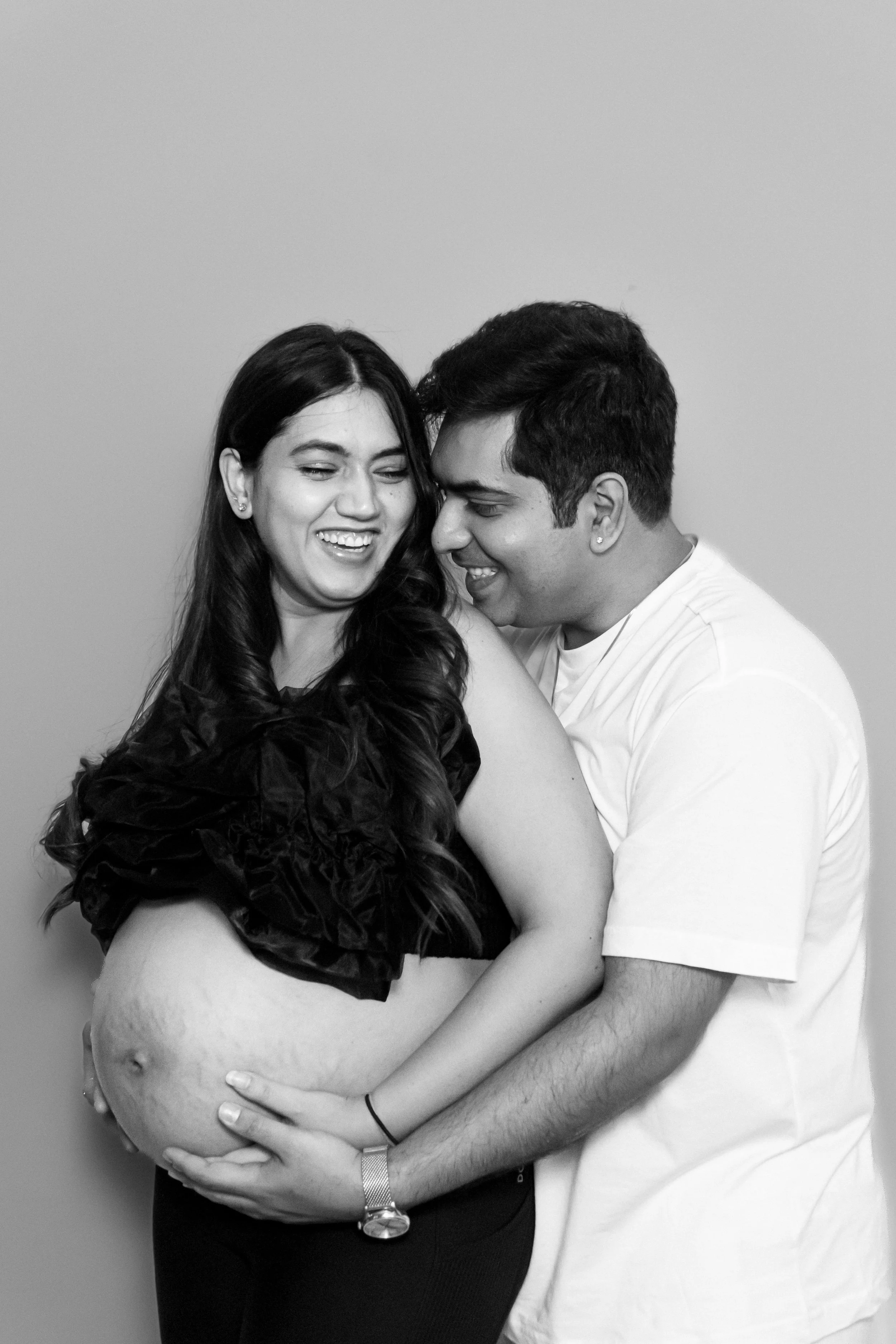 Black and white photo of a pregnant woman holding her belly, smiling, with a man embracing her from behind, both smiling and close to each other.
