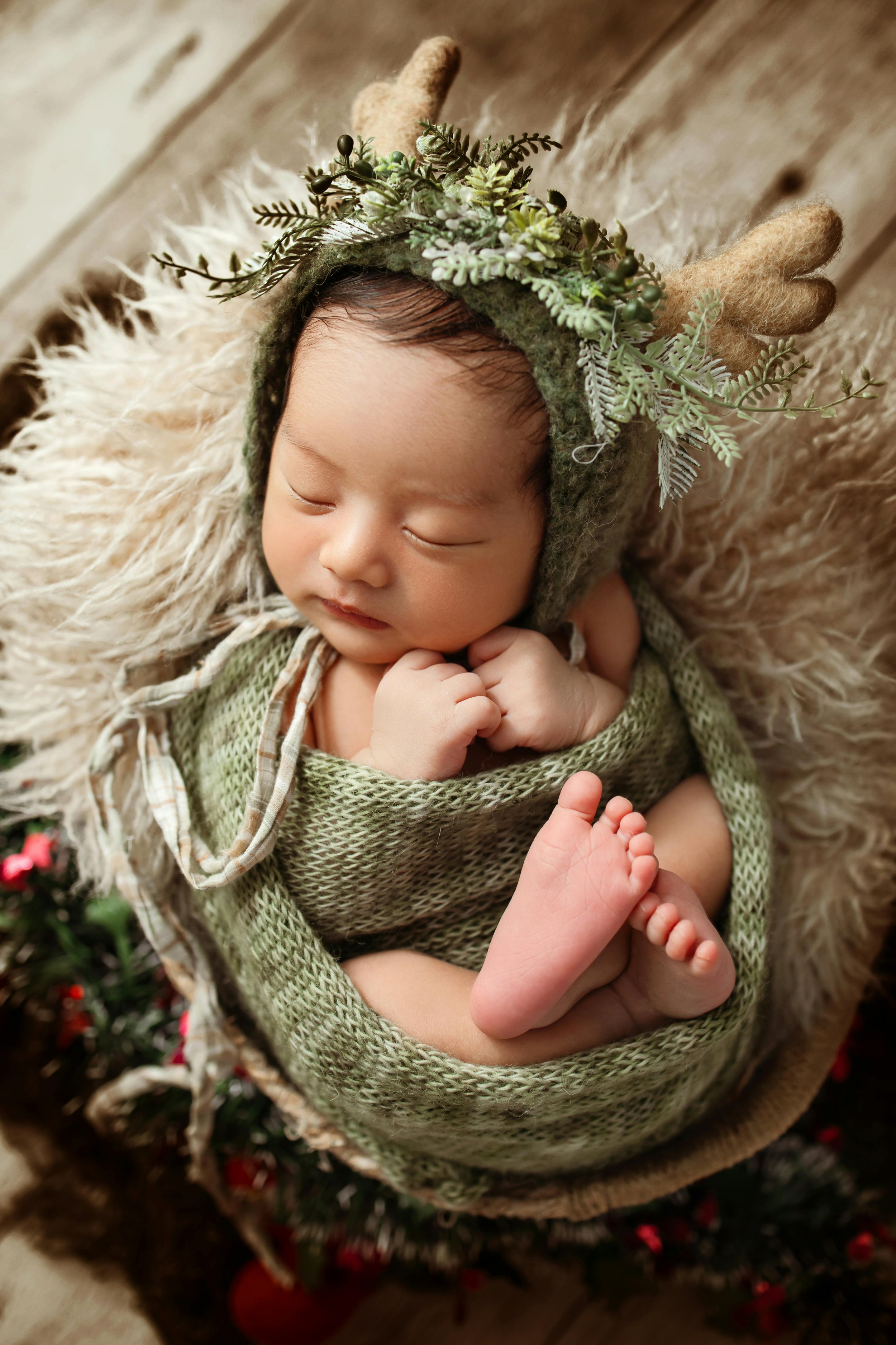 A sleeping baby wearing a green knitted outfit and a hat decorated with greenery and antlers, lying on a fluffy cream-colored blanket.