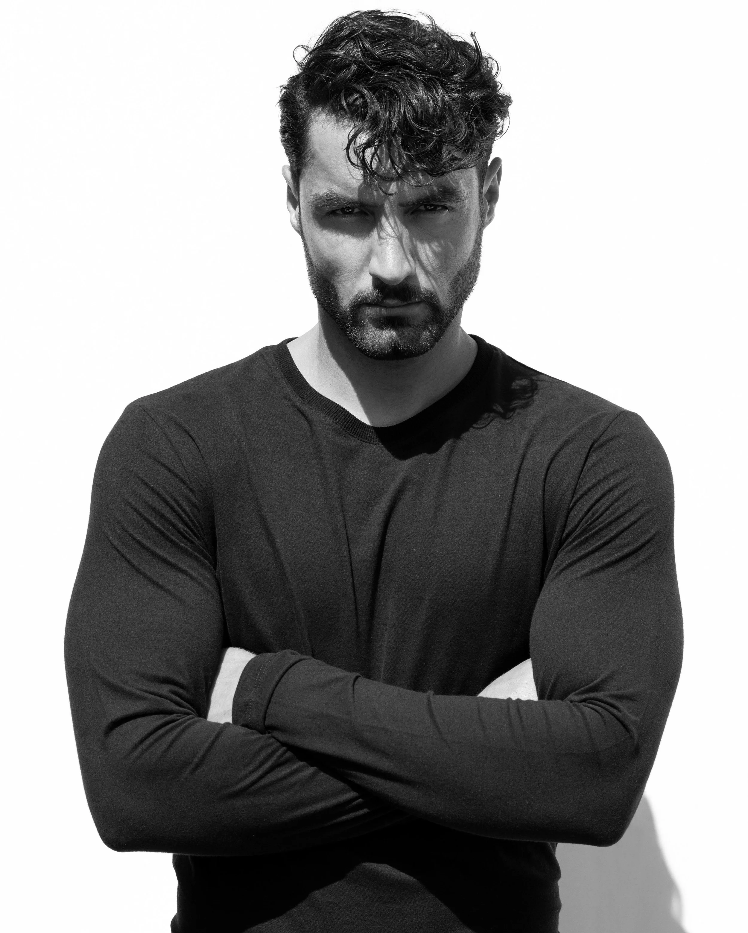 Black and white photo of a man with curly hair and a beard, crossing his arms and staring intensely at the camera against a plain background.