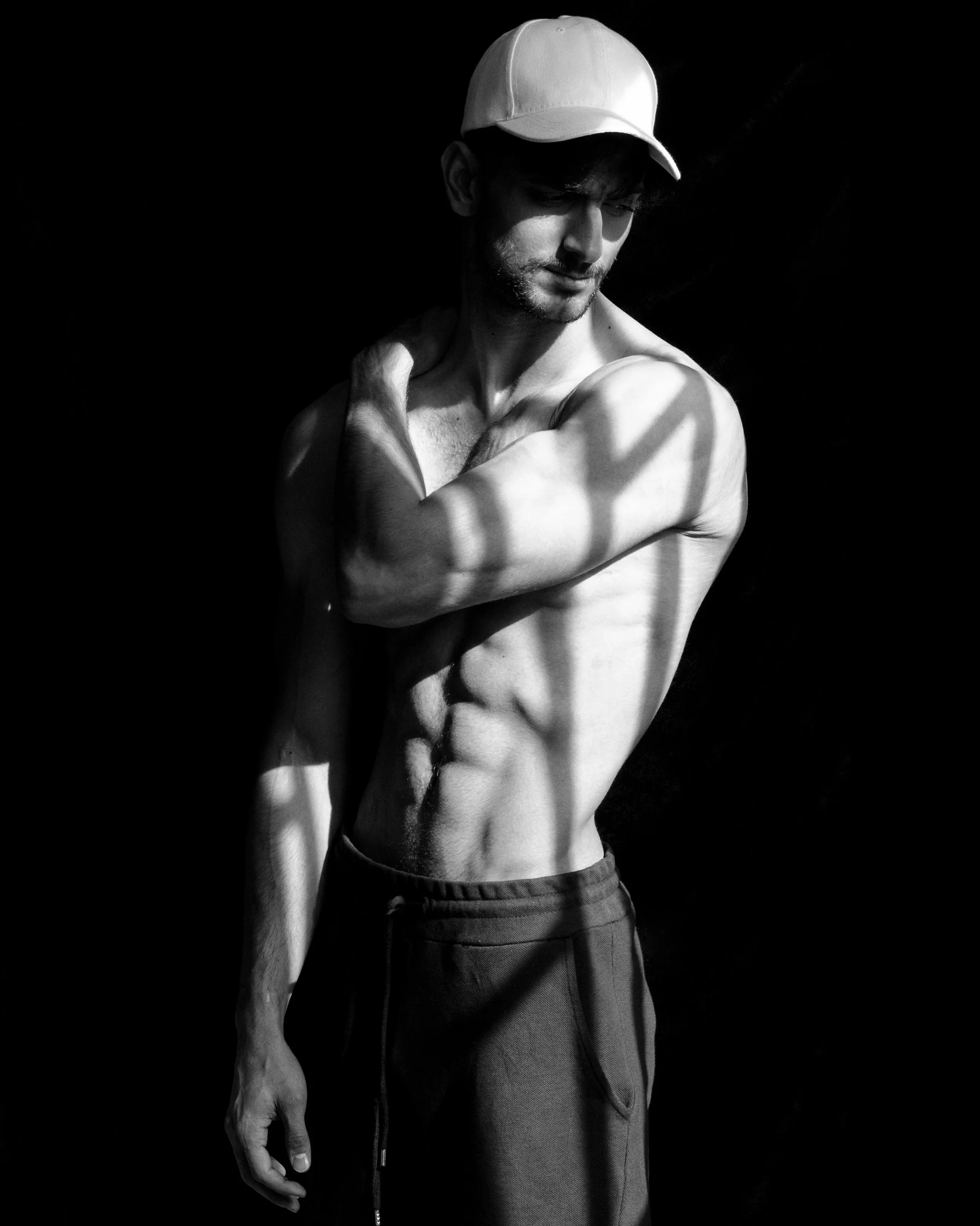 A black-and-white photograph of a shirtless young man wearing a white cap, standing against a dark background. Shadows cast across his body create a pattern of light and shadow.
