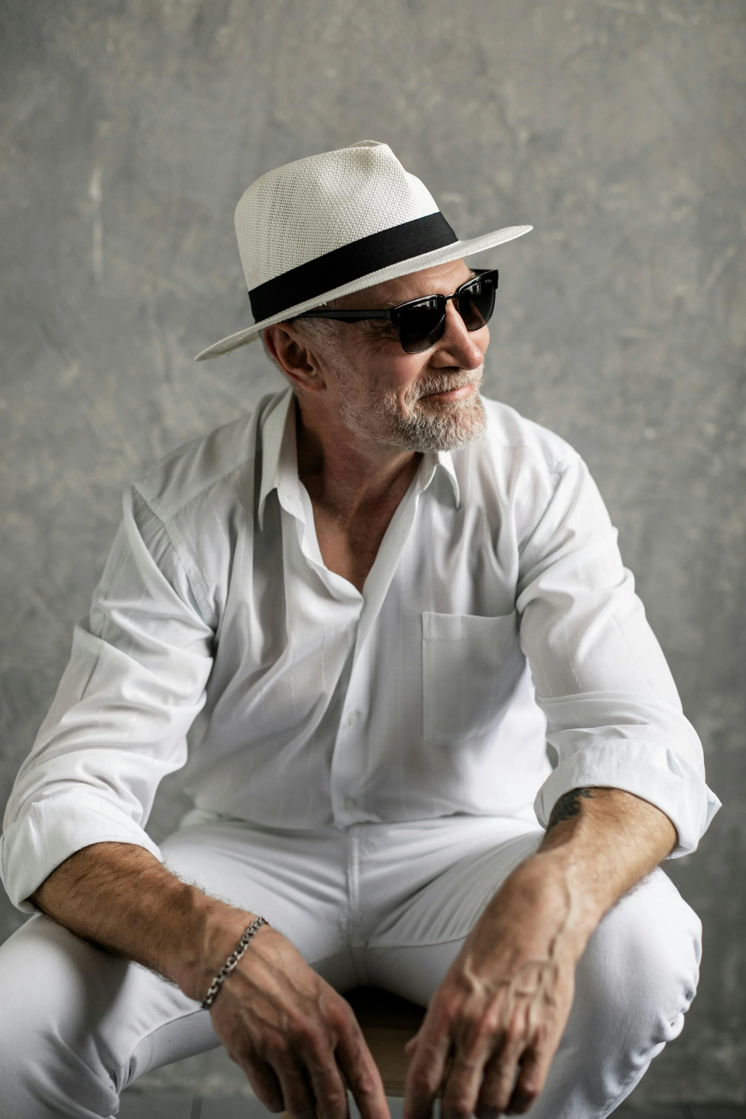 A man wearing a white fedora, black sunglasses, white shirt, and white pants, sitting against a textured gray background.