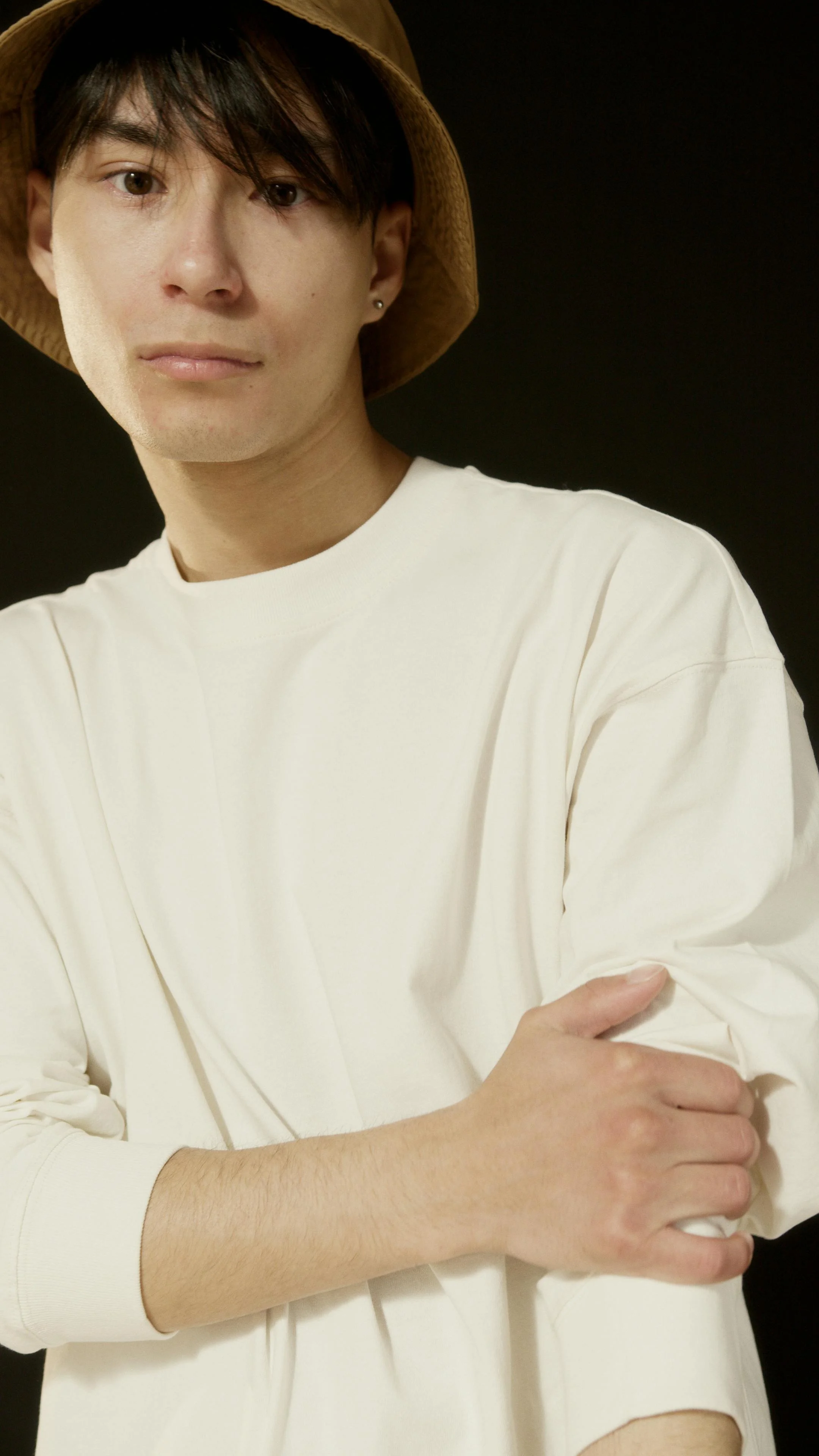 A young man wearing a brown straw hat and a white long-sleeve shirt, standing against a plain black background.