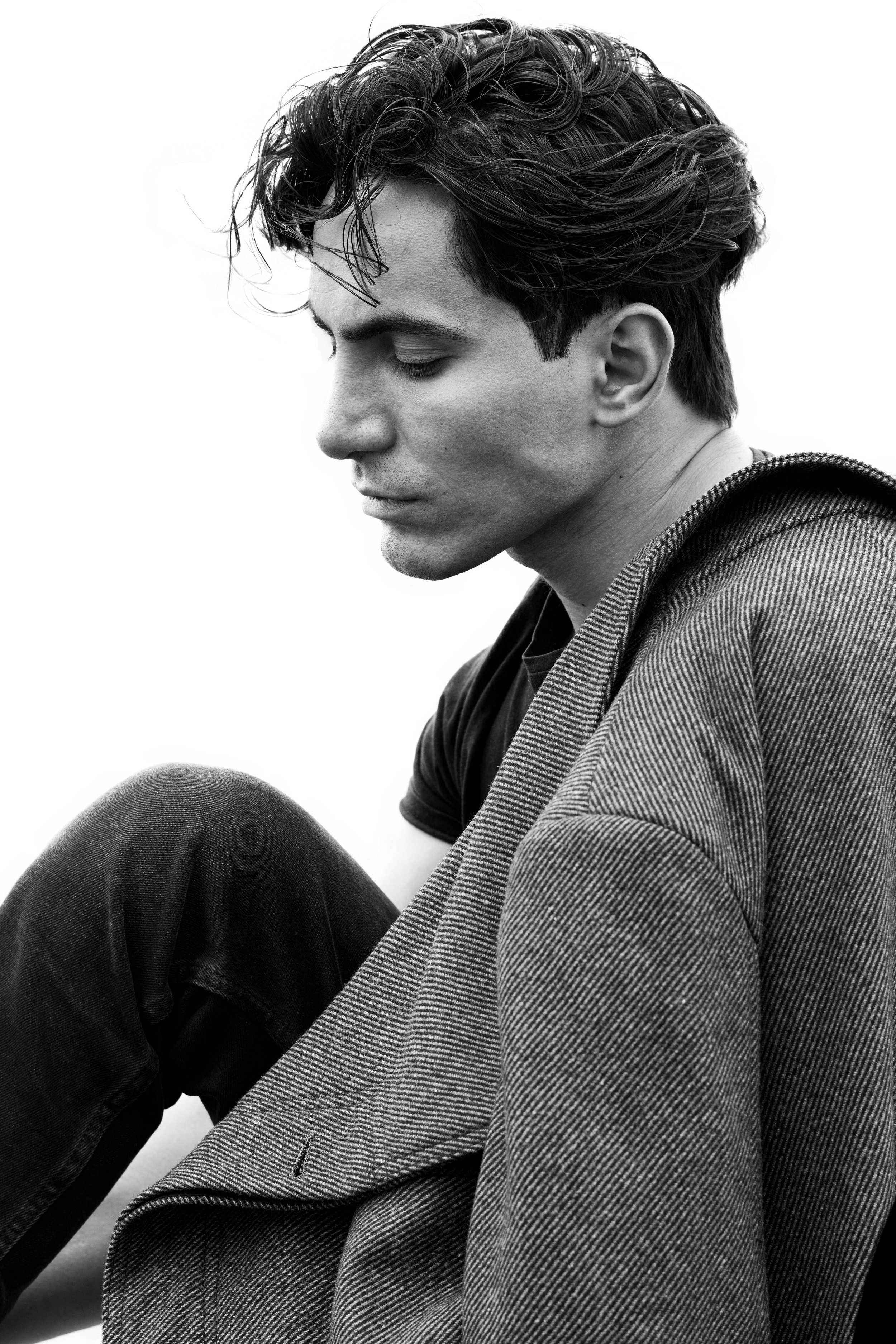A black and white photo of a young man with disheveled hair, sitting with his head tilted downward, eyes closed, wearing a dark t-shirt and a textured blazer.