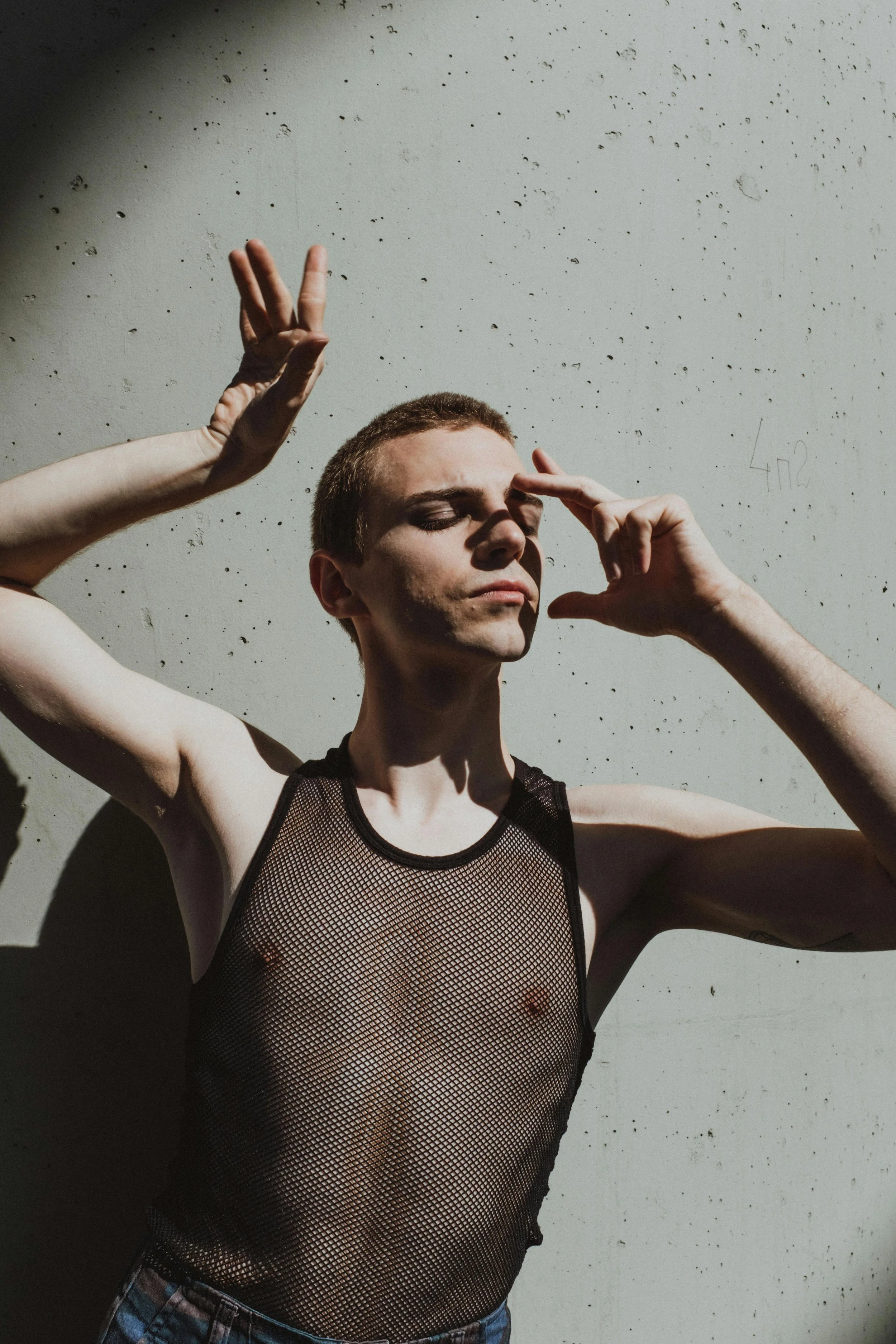 Person with short hair wearing a black fishnet tank top standing against a textured gray wall with sunlight casting shadows.
