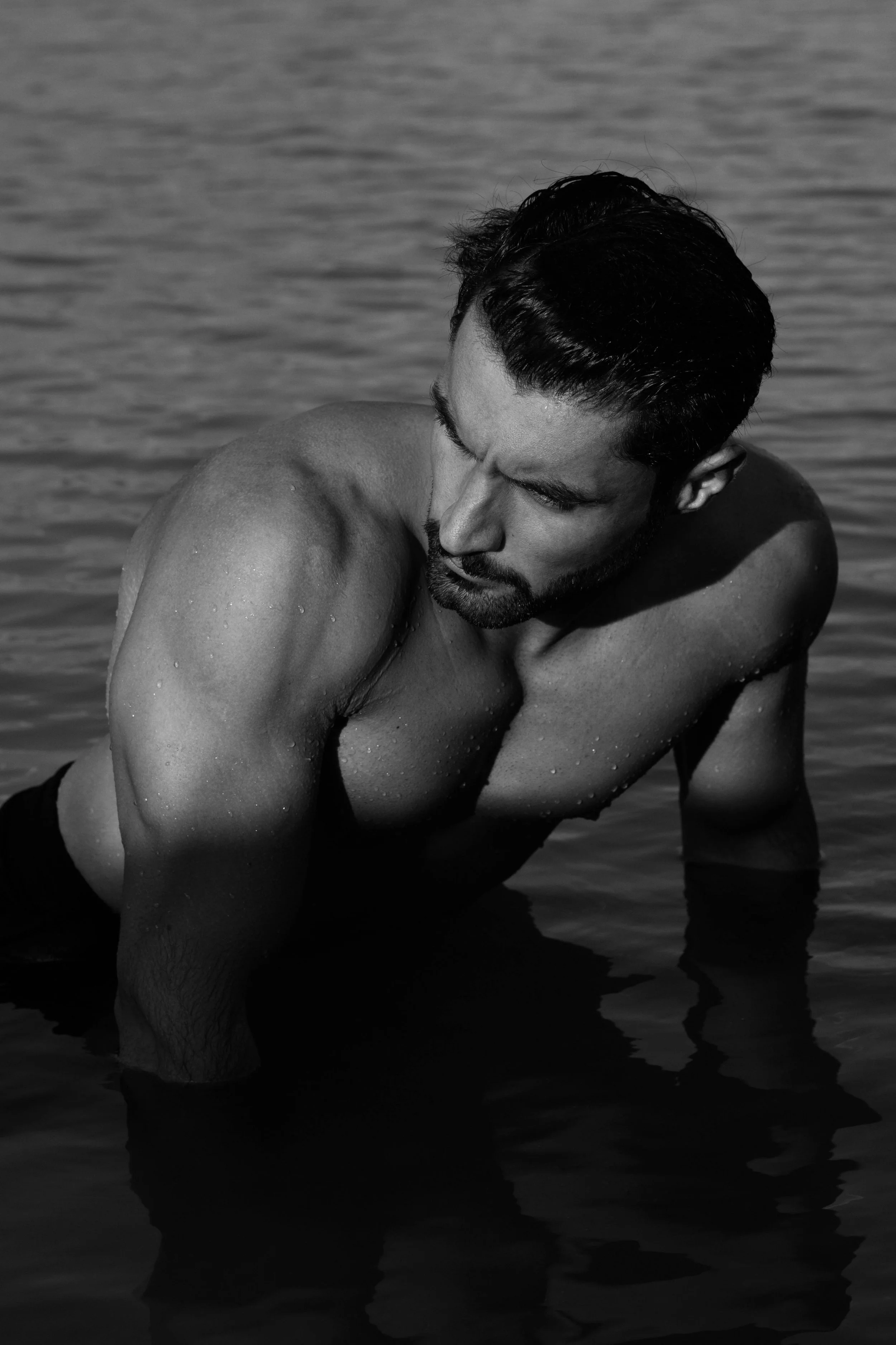 A black and white photo of a muscular man with dark hair, sitting in water with droplets on his skin, looking downward.