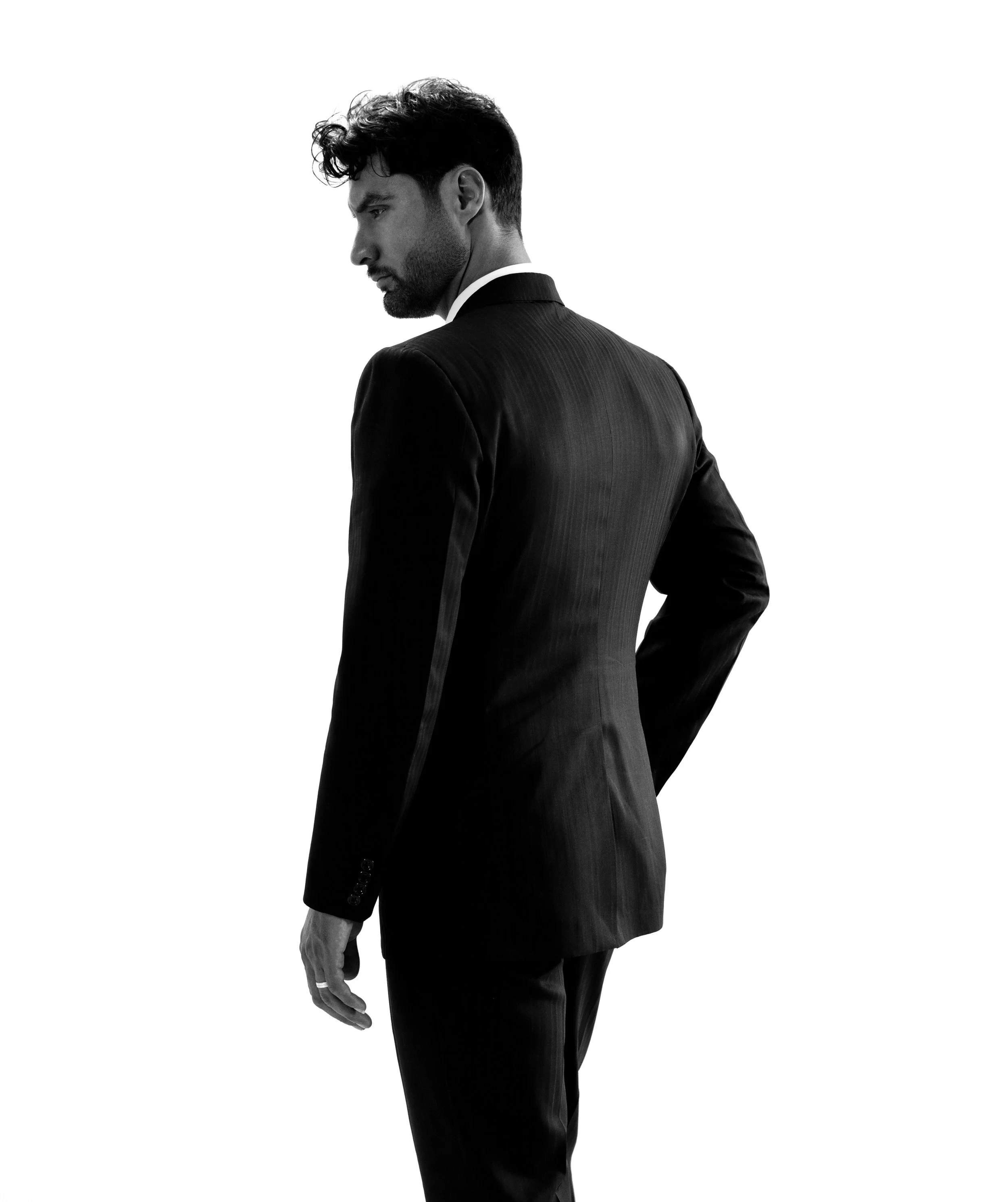 A black and white photo of a man in a suit standing sideways, looking downward, with a plain white background.