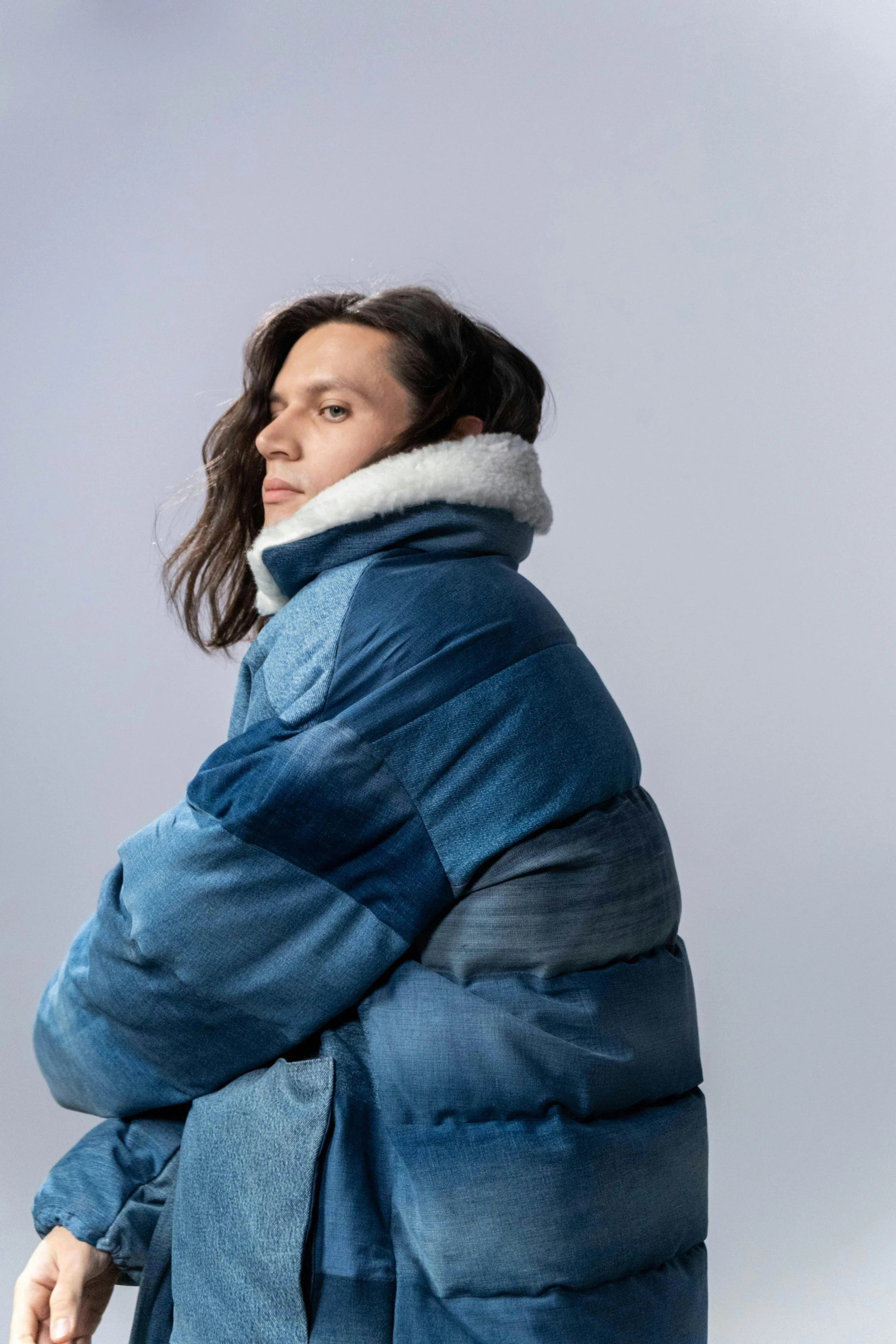 A person with shoulder-length dark hair looking over their shoulder, wearing a blue and gray puffer jacket with a white fleece-lined collar, against a plain light background.