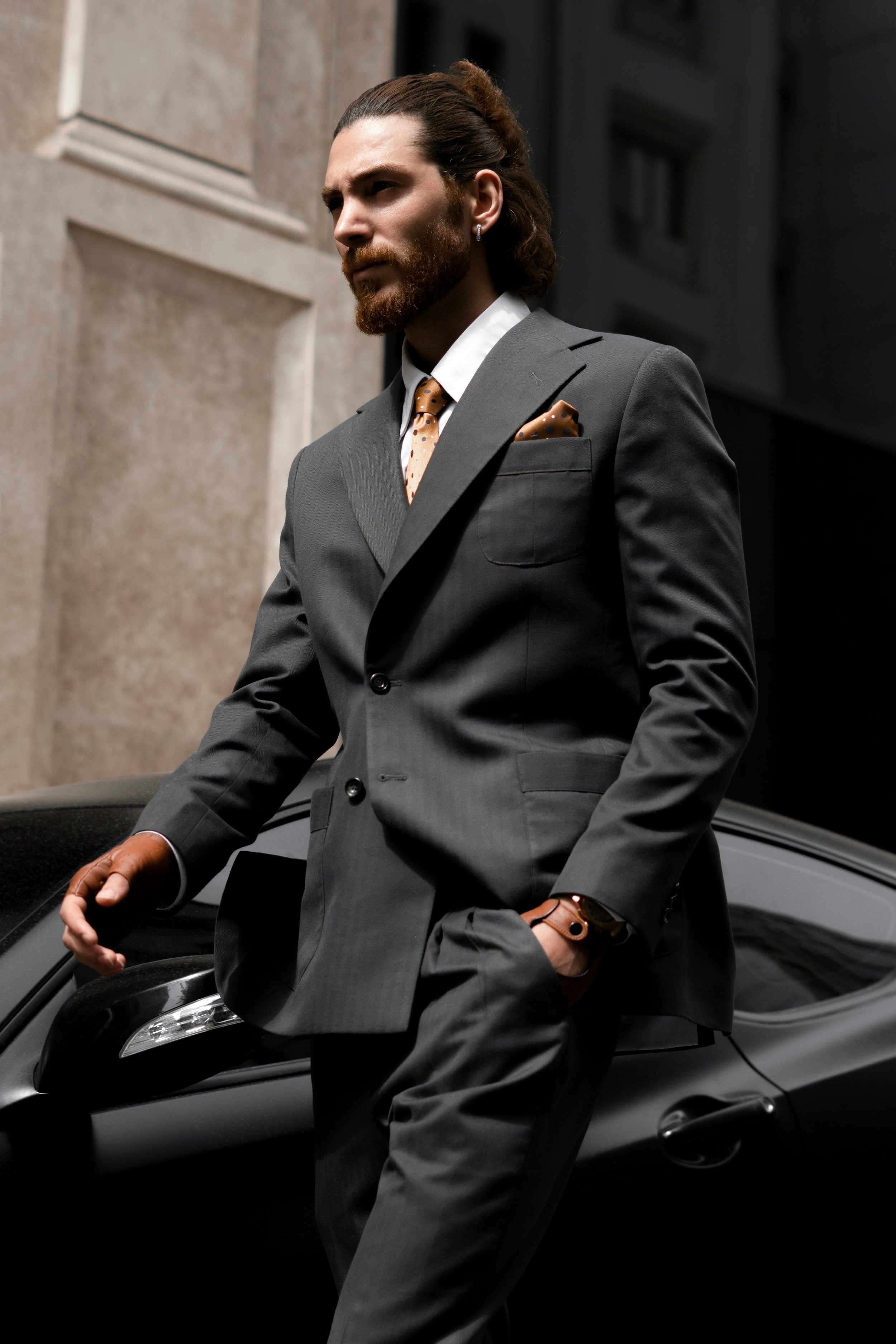 A man in a gray suit with a patterned tie and pocket square leaning against a black sports car in an urban setting.