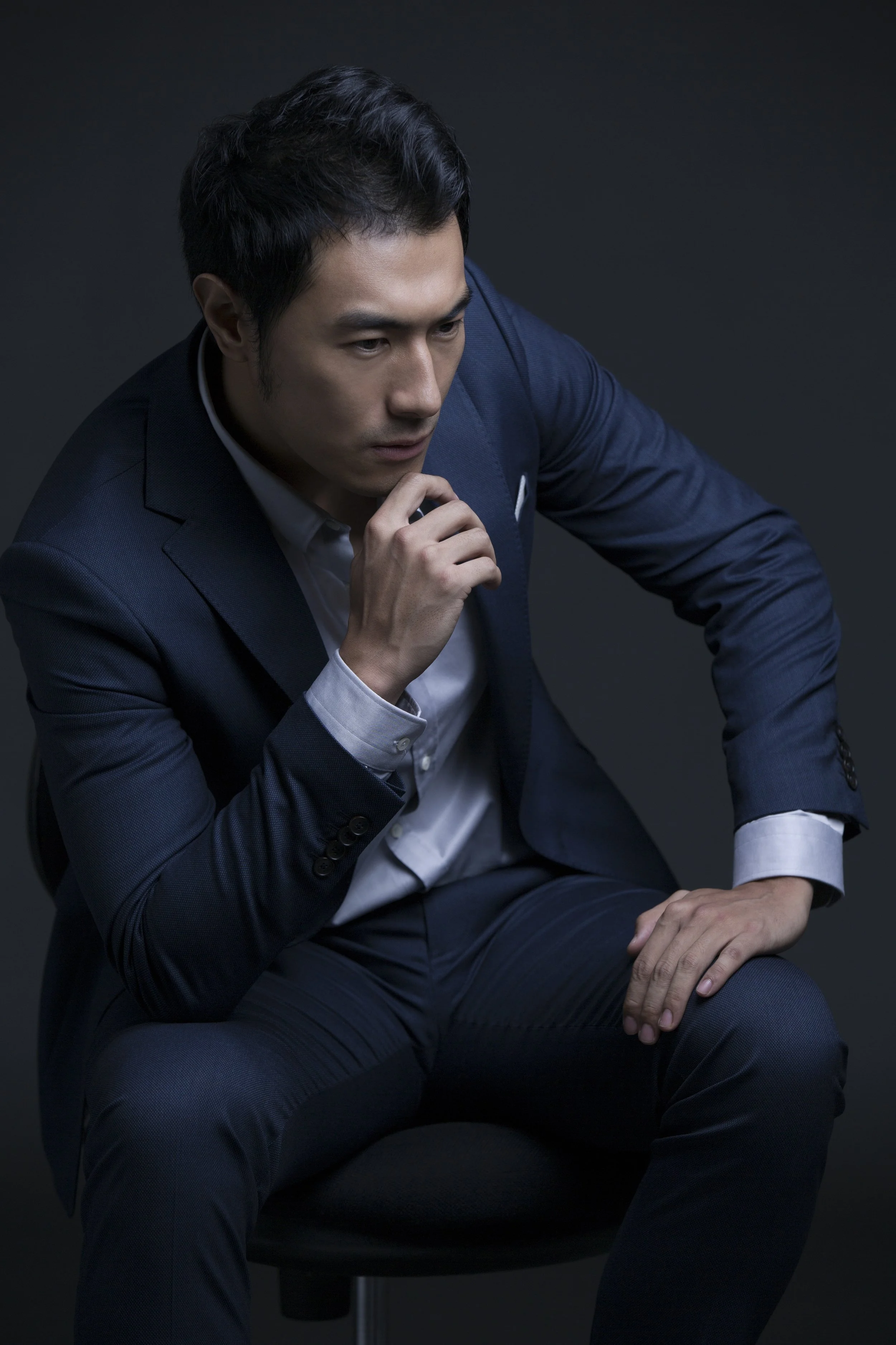 A man dressed in a dark blue suit with a white shirt, seated on a black chair, in a thoughtful pose with his hand near his chin, against a dark background.