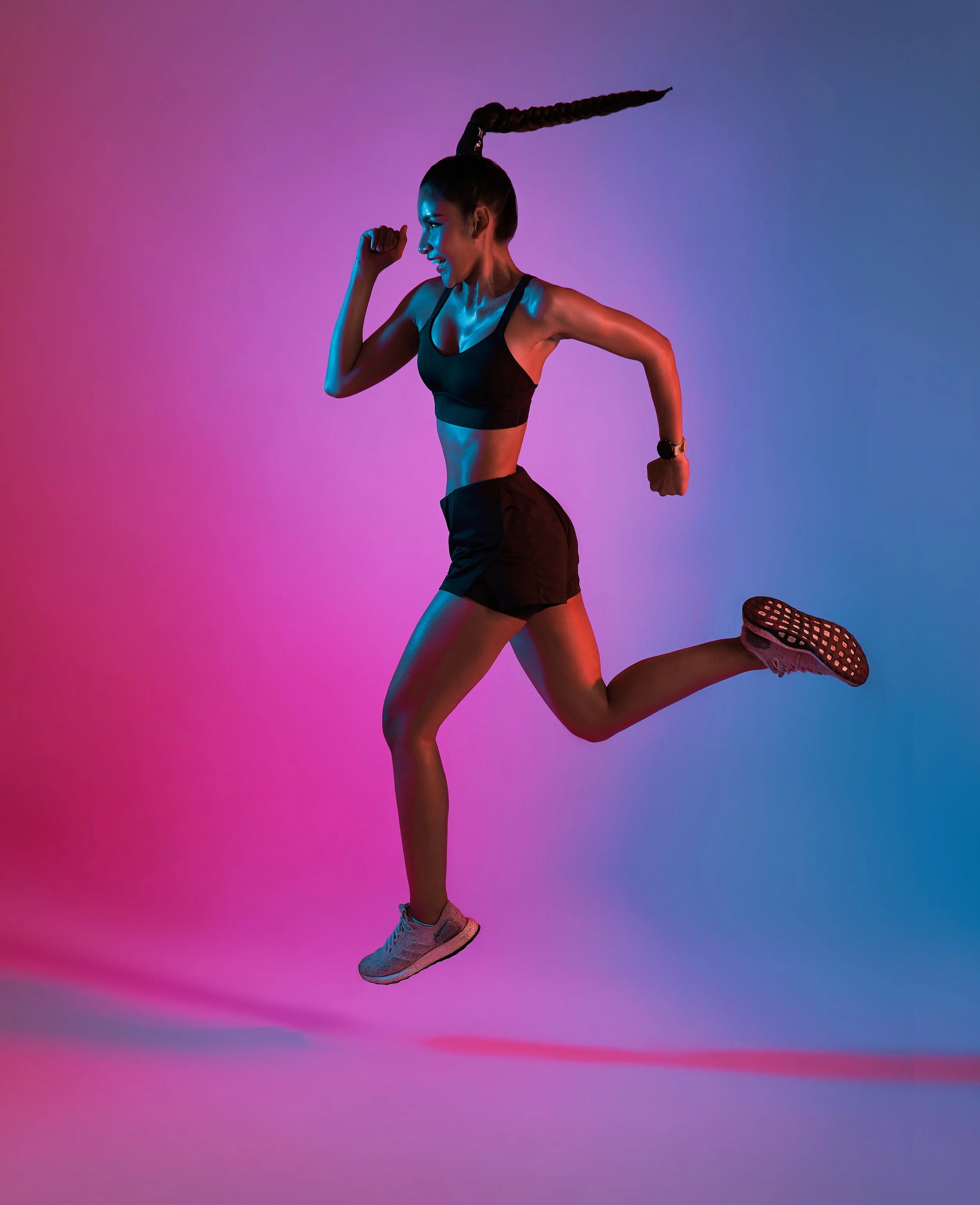 A woman with a long braid, wearing workout clothes, is mid-air in a jump against a colorful pink and blue gradient background.