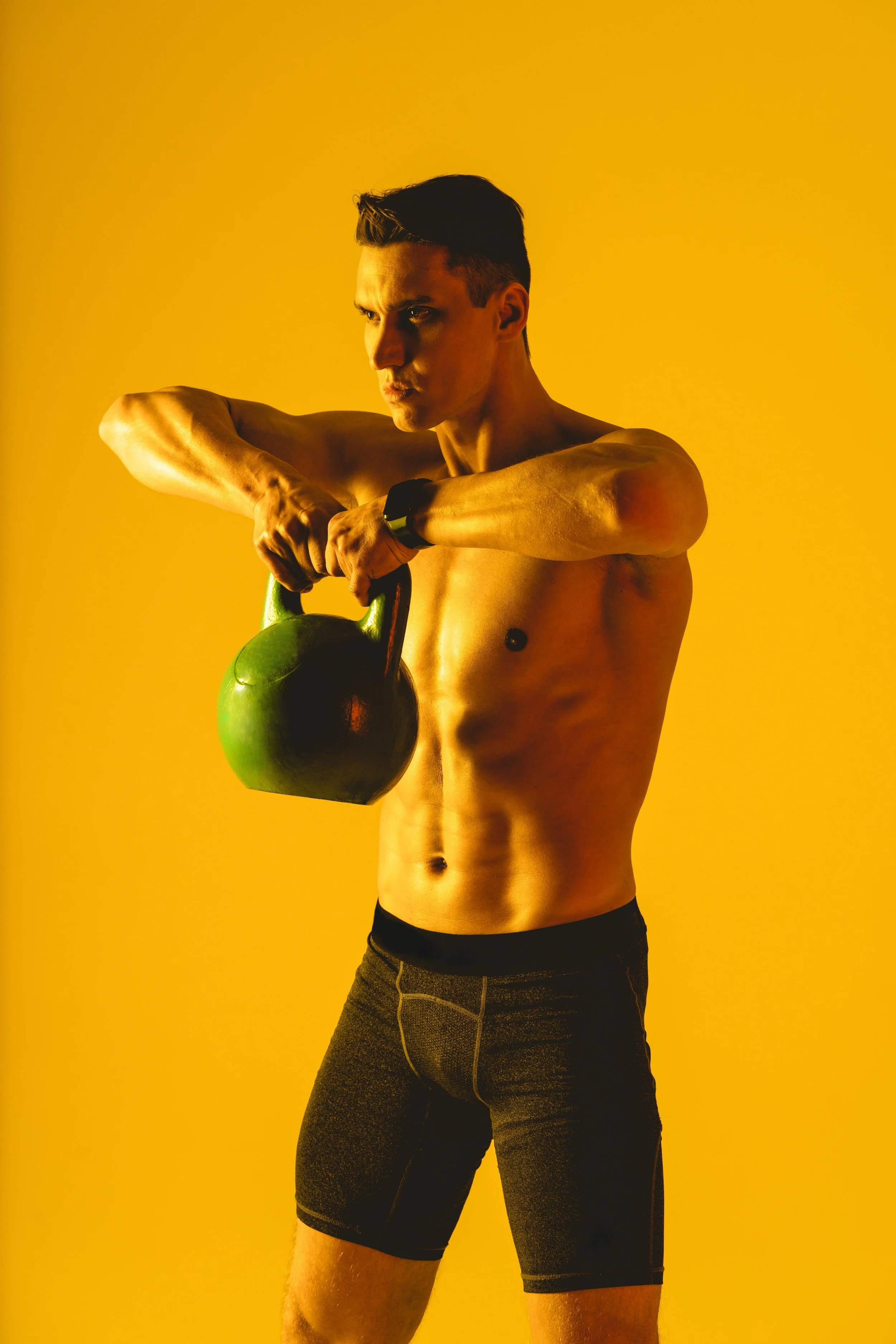 A shirtless man with a fitness watch on his wrist holds a black kettlebell against a yellow background.