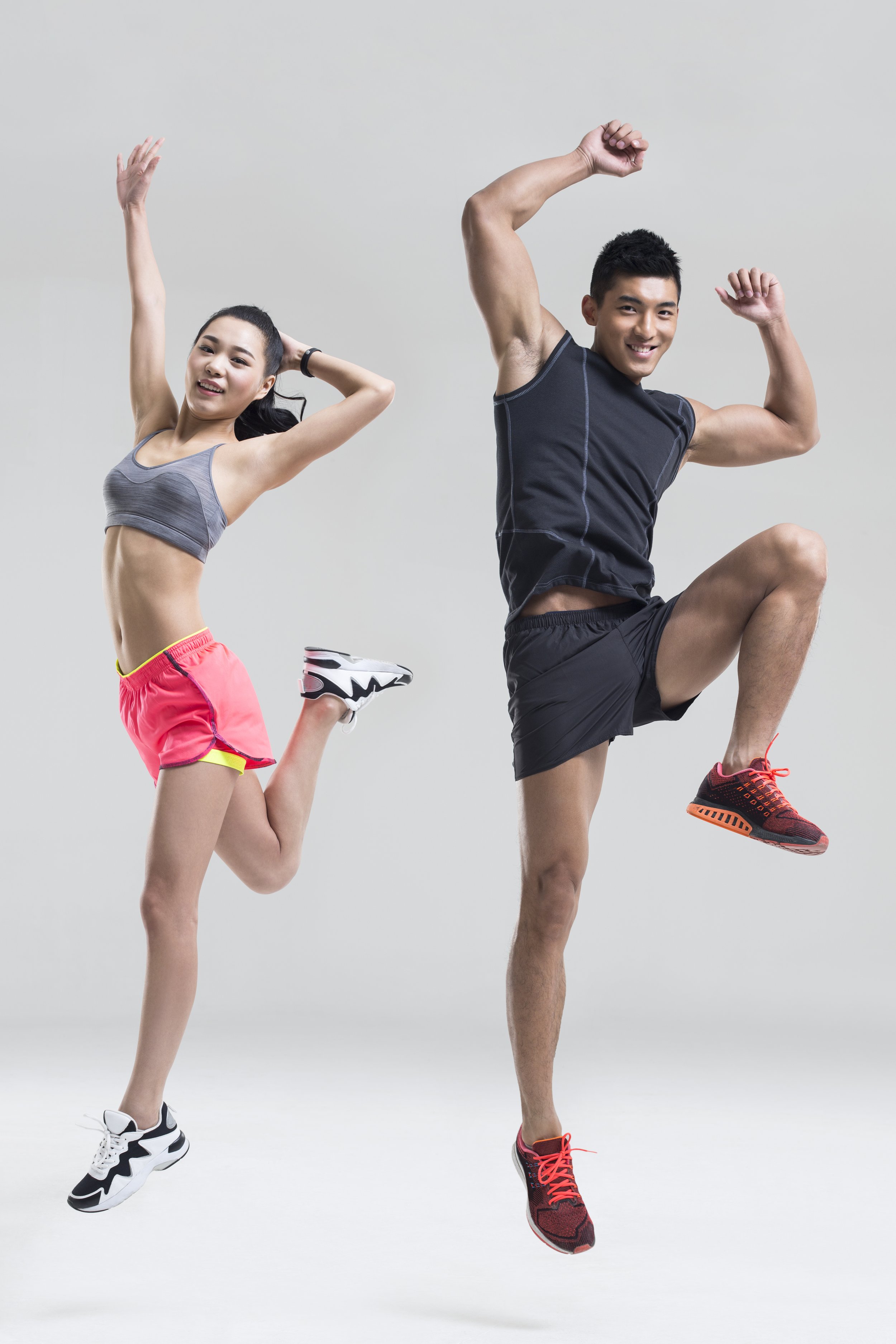 Two fit young people, a woman and a man, jumping in athletic clothes against a plain background, smiling and looking at the camera.