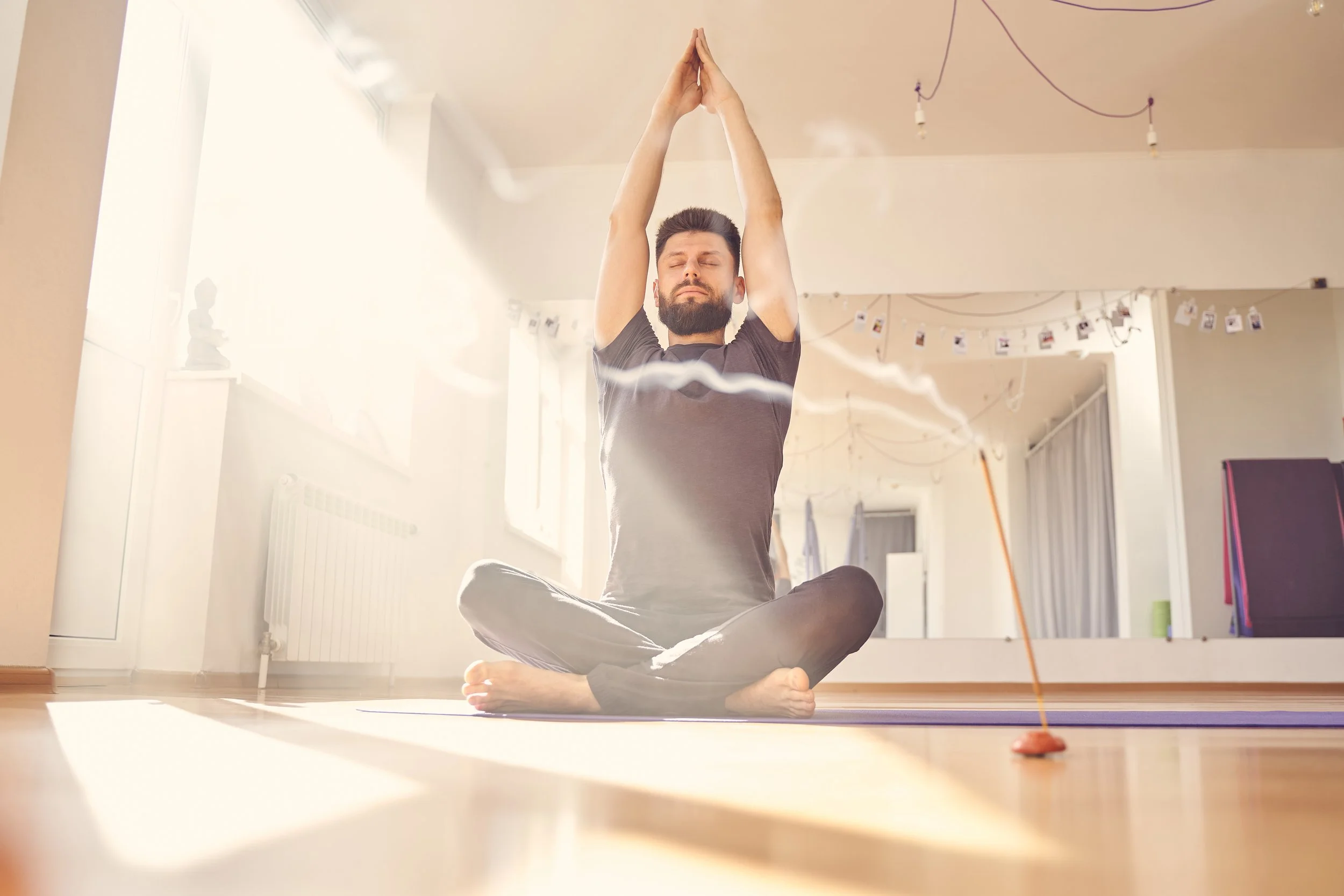 Man practicing meditation on a purple yoga mat in a bright room with sunlight, sitting cross-legged with hands in prayer pose above his head, surrounded by decorative string lights and personal photos.