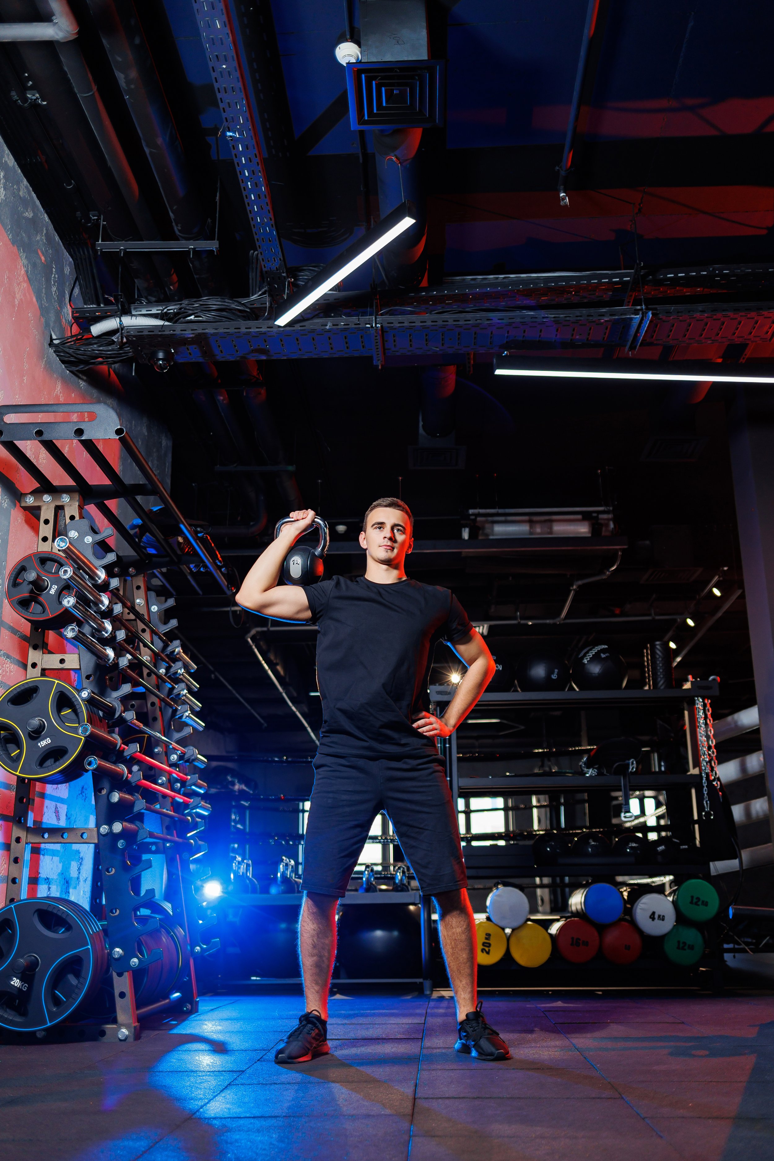 A young man in workout clothes holding a kettlebell in a gym with weights and workout equipment in the background.