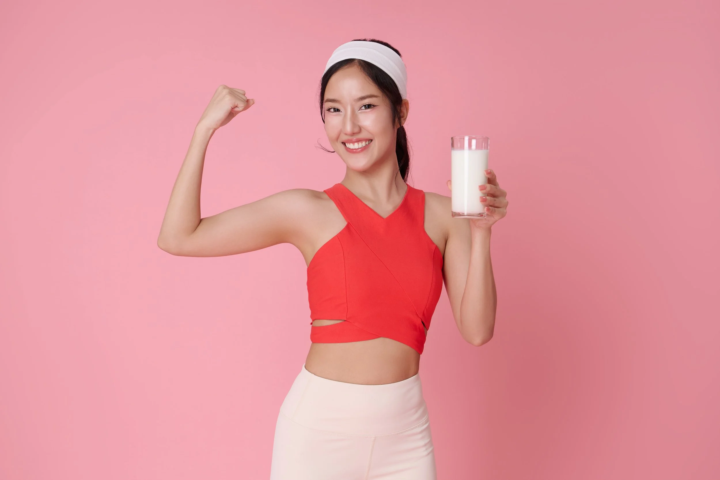 Young woman in workout clothes holding a glass of milk and flexing her arm on pink background.