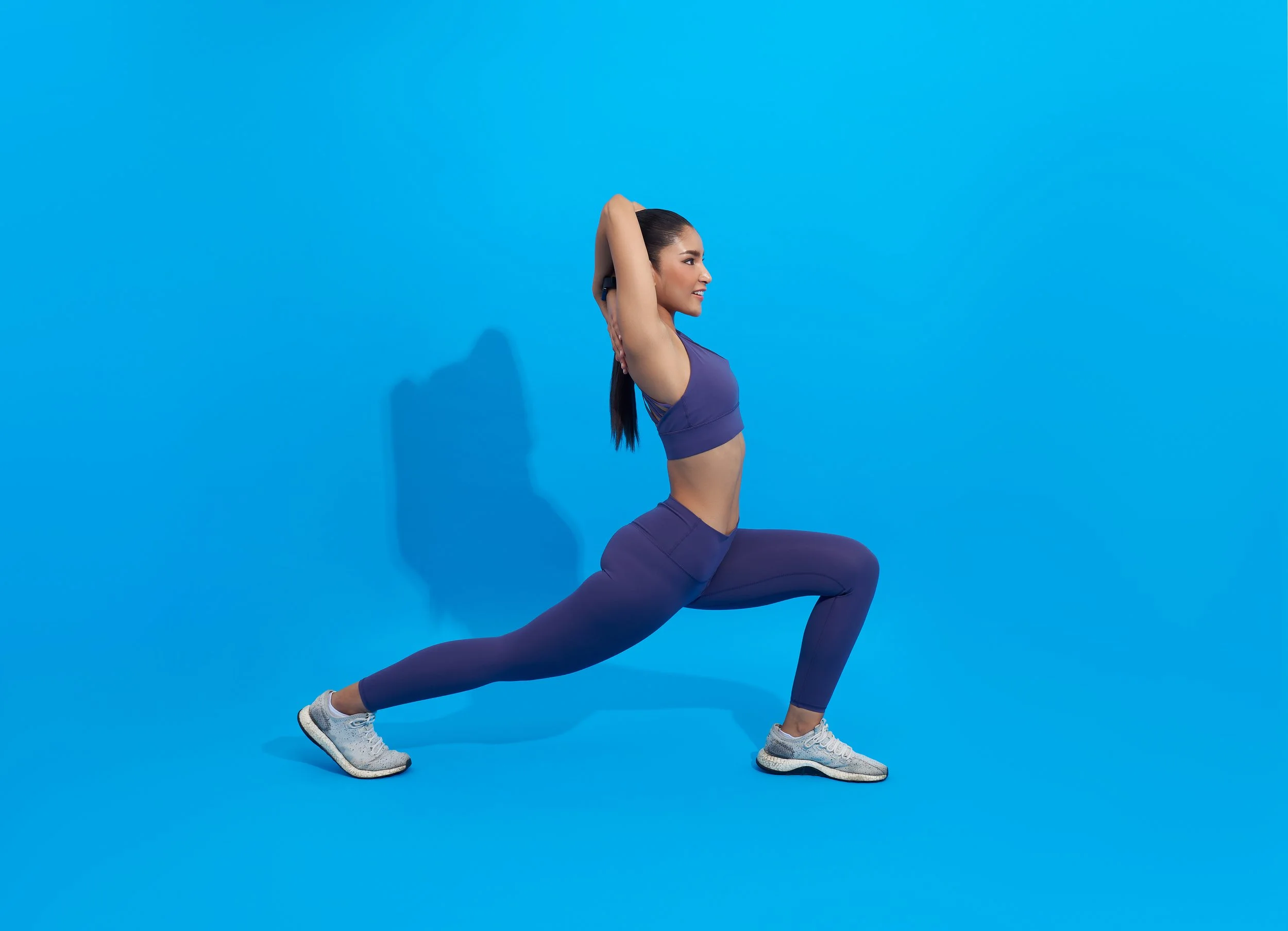 A woman in athletic wear performing a yoga lunge pose against a solid blue background.