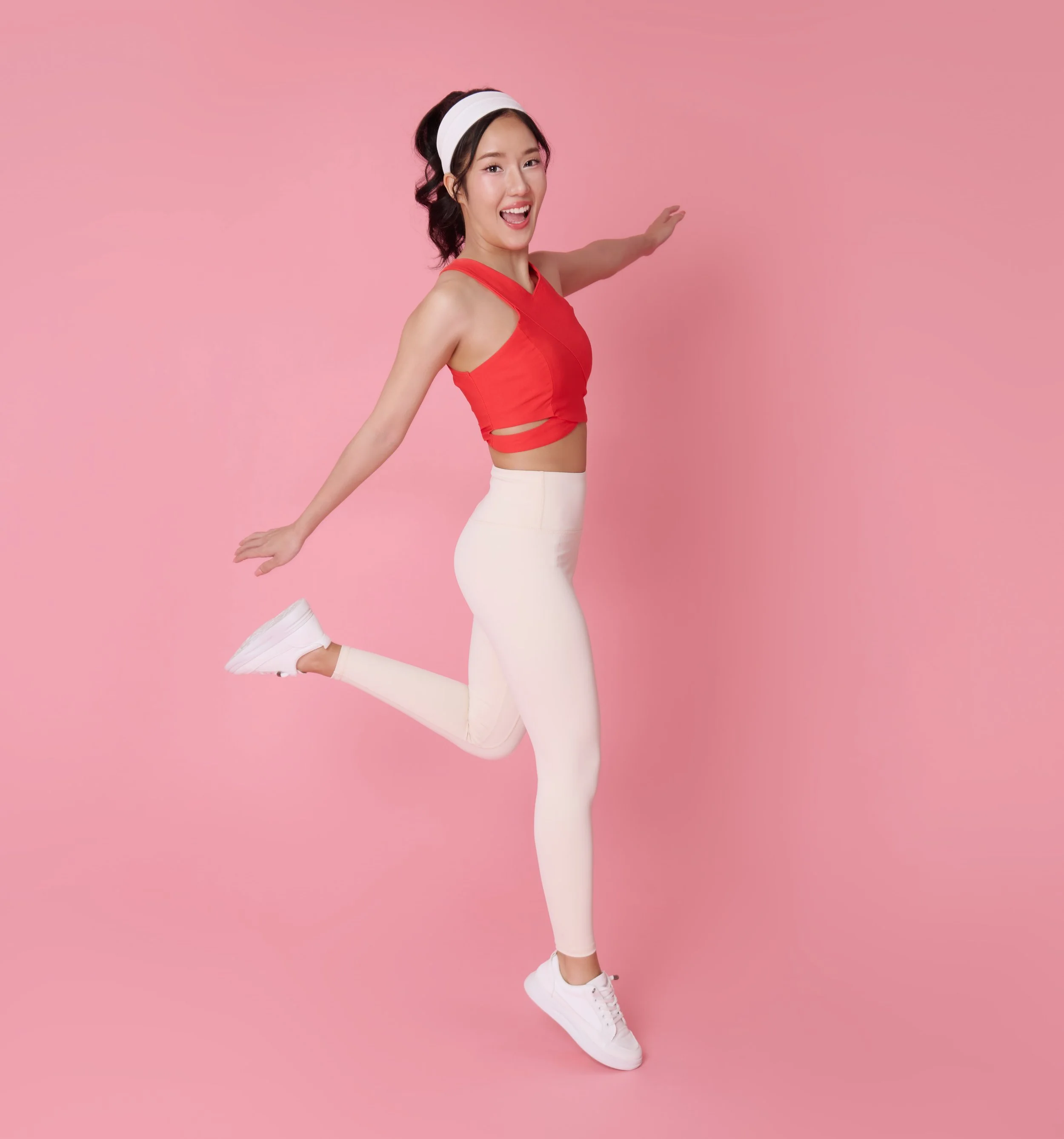 A woman in a red crop top and white leggings jumping against a pink background, smiling and with arms extended outward.