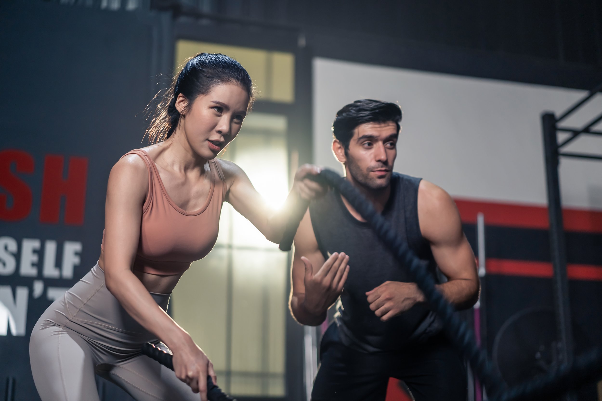 A woman and a man working out together in a gym using battle ropes, with the woman demonstrating proper form and the man following her lead, both focused and engaged in training.