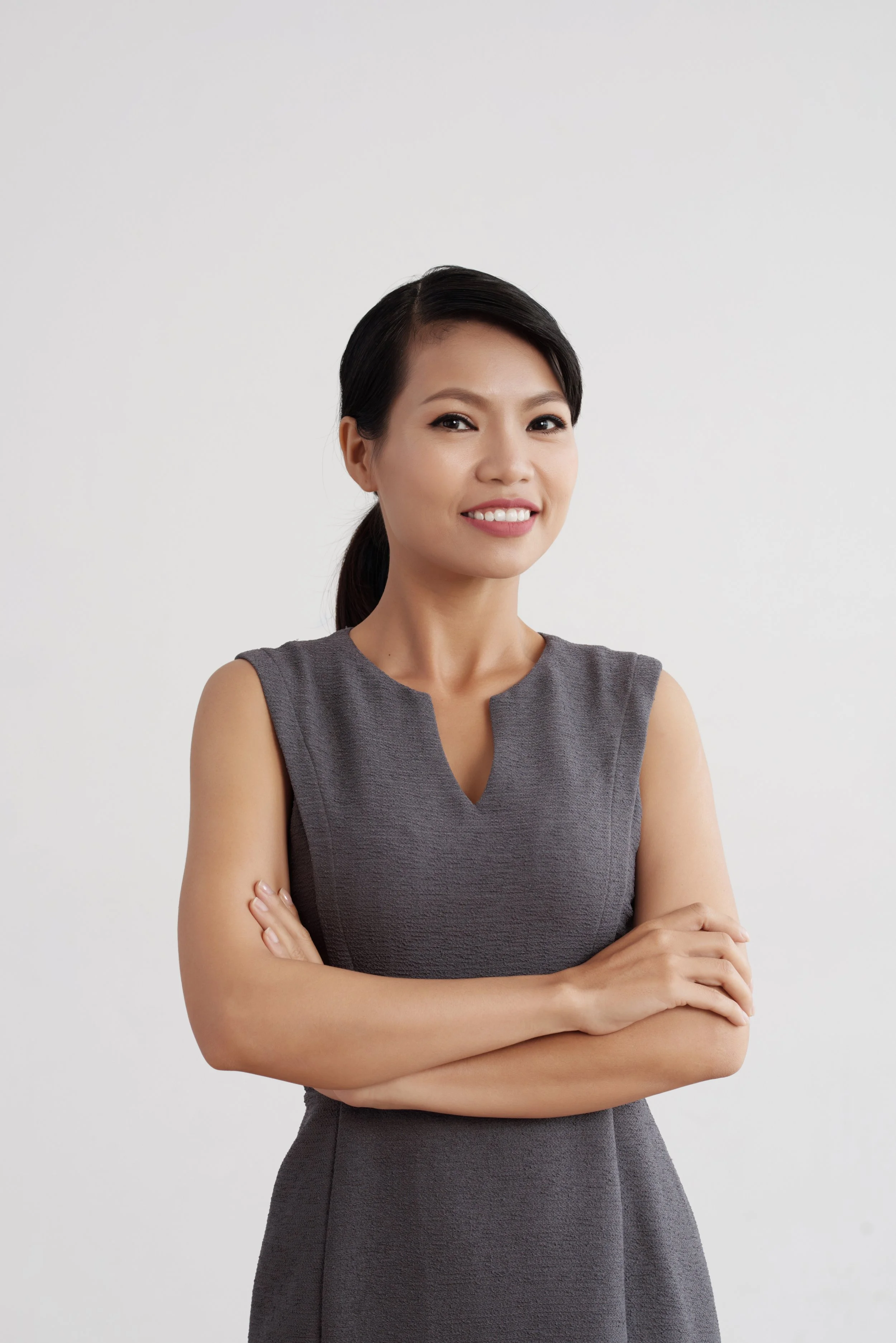 Portrait of a young woman with dark hair in a ponytail, smiling, wearing a sleeveless gray dress, standing against a plain white background.