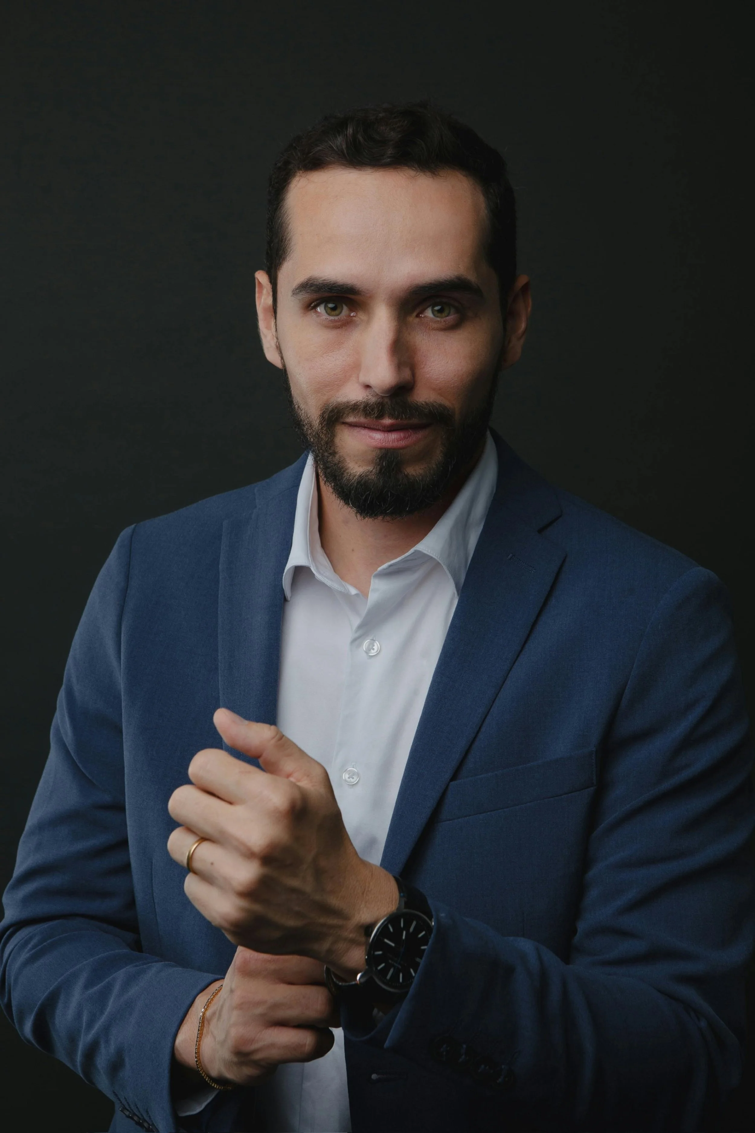 A man with a well-groomed beard and short dark hair wearing a dark blue suit, white shirt, and a black watch, posing against a dark background.