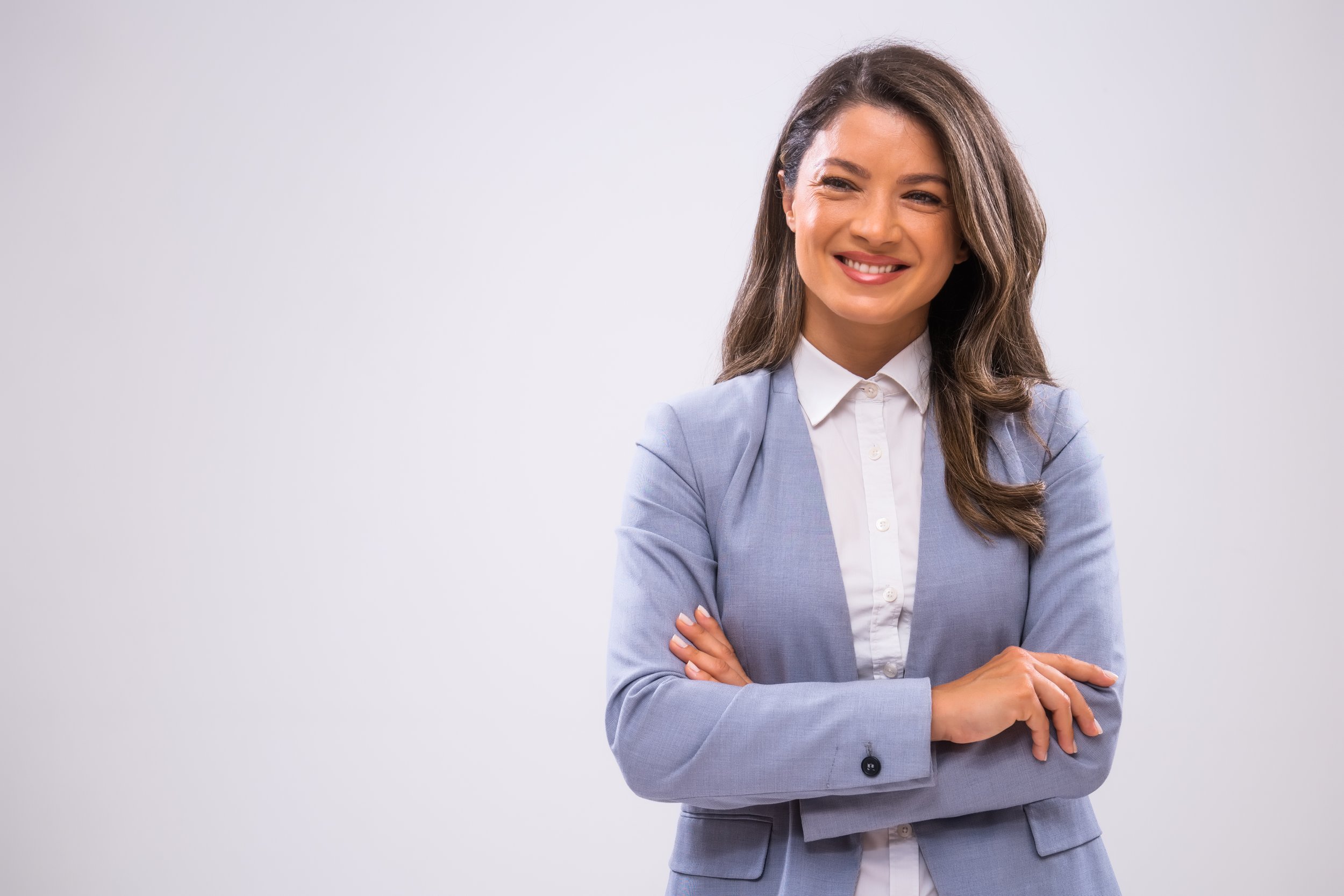 A woman with long, wavy brown hair, wearing a light gray blazer and white shirt, smiling with her arms crossed against a plain light gray background.