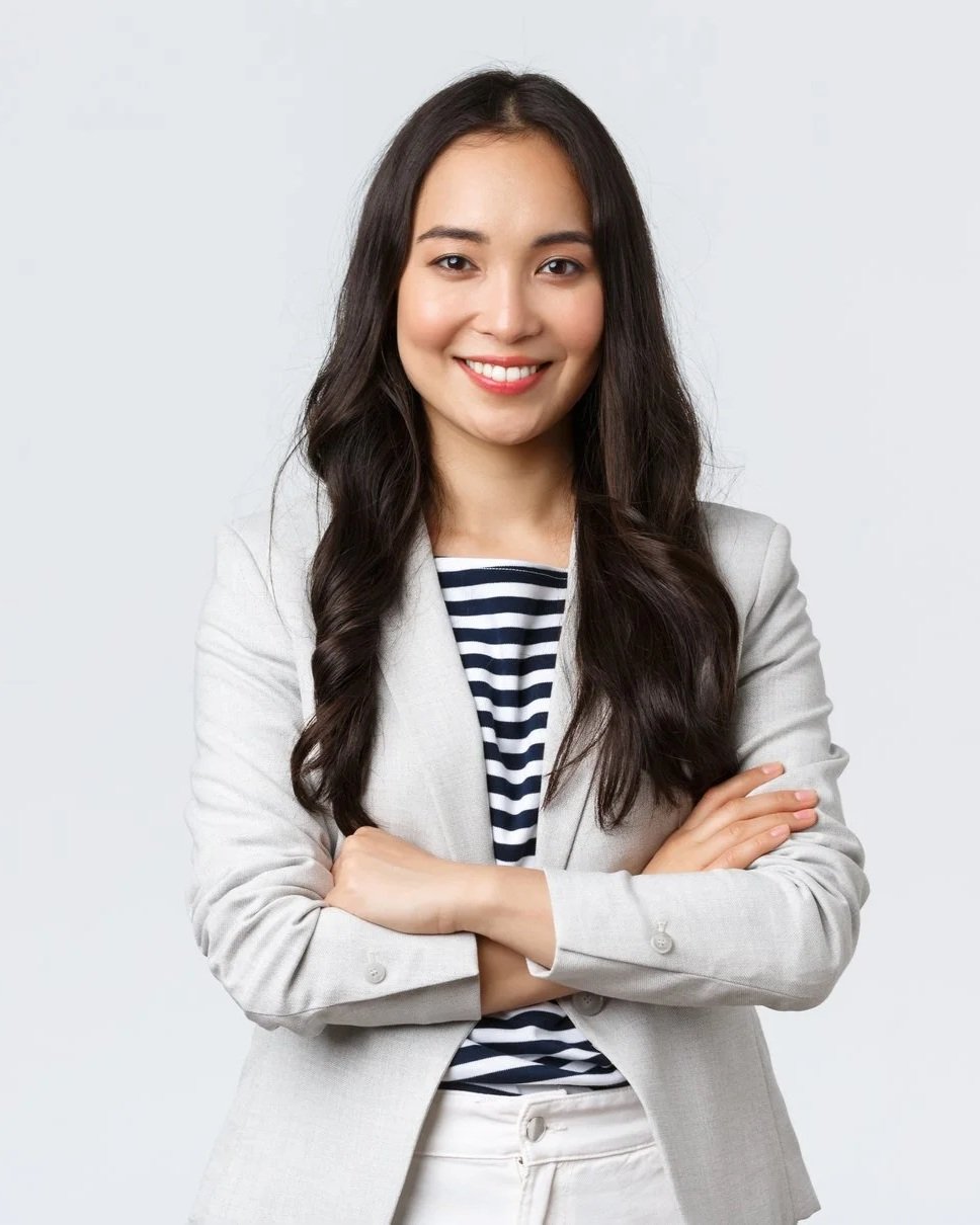 A young woman with long dark hair smiling, dressed in a light blazer over a striped shirt, with arms crossed, standing against a plain light background.