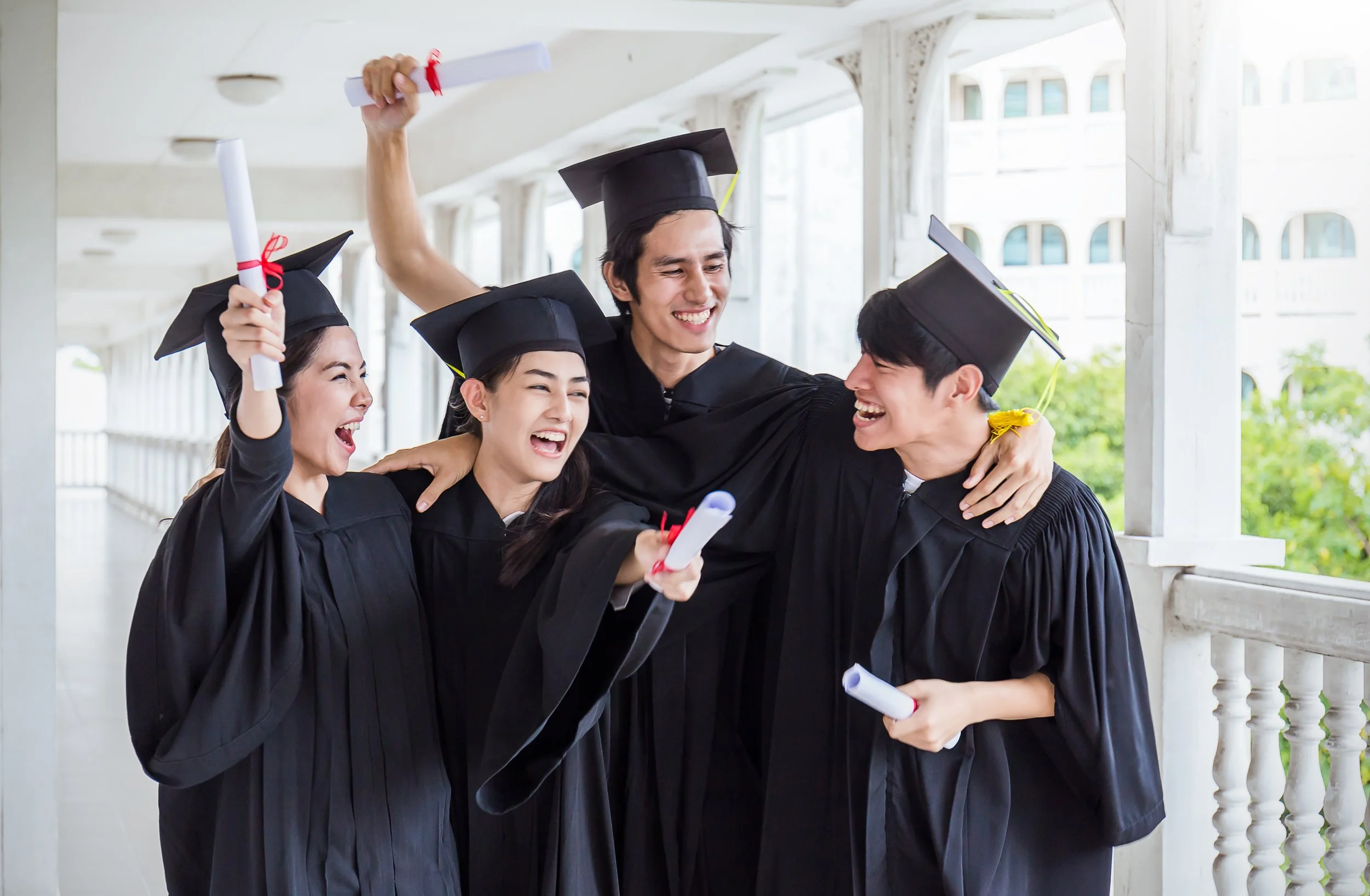 Group of four diverse graduates in black caps and gowns celebrating and hugging each other in a corridor outside, holding diplomas.