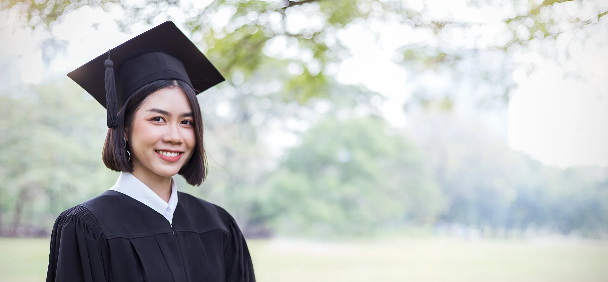 Young woman in a graduation cap and gown smiling outdoors with blurred trees in the background.