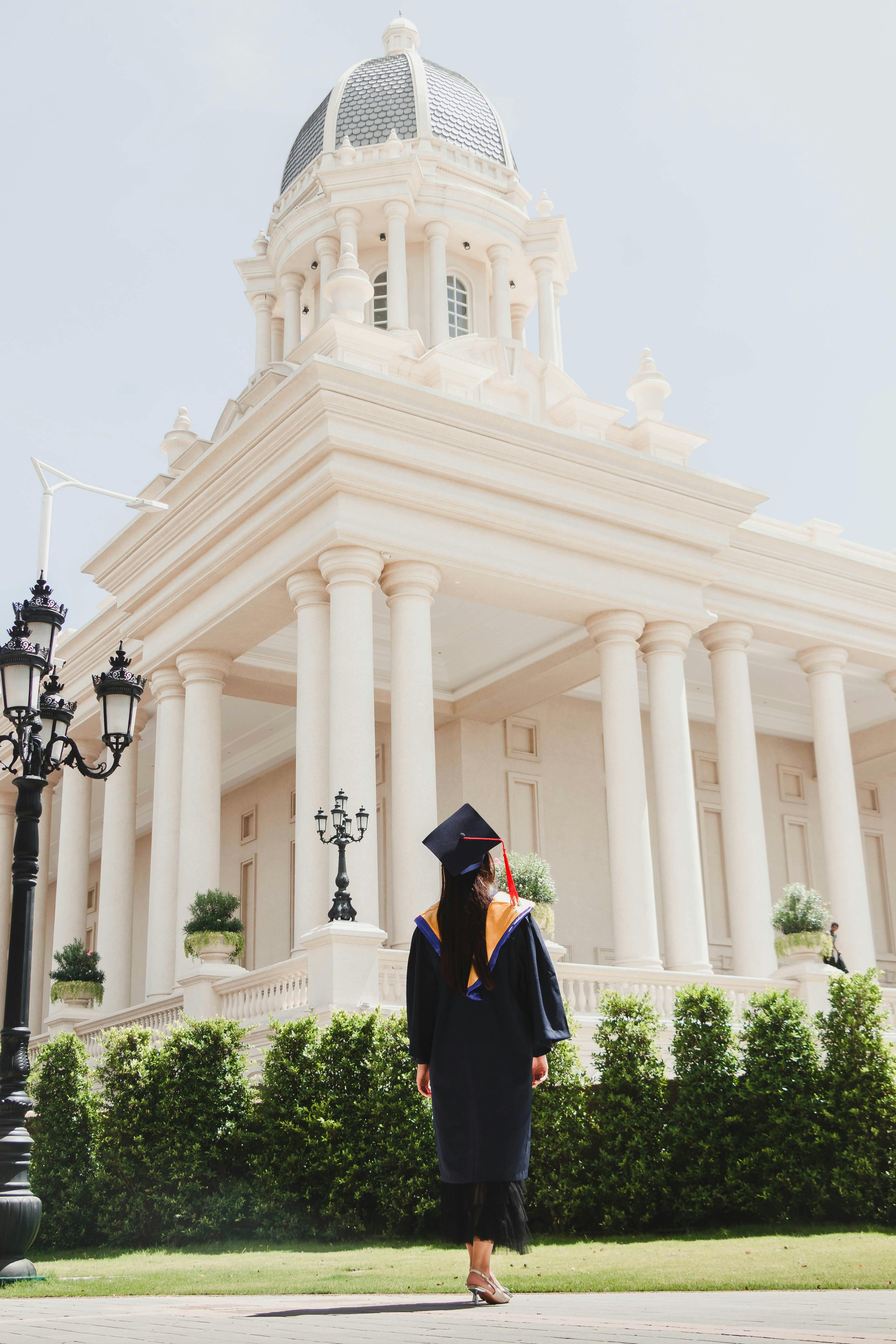 A person in a graduation gown and cap stands facing a large, white classical-style building with tall columns and a dome.