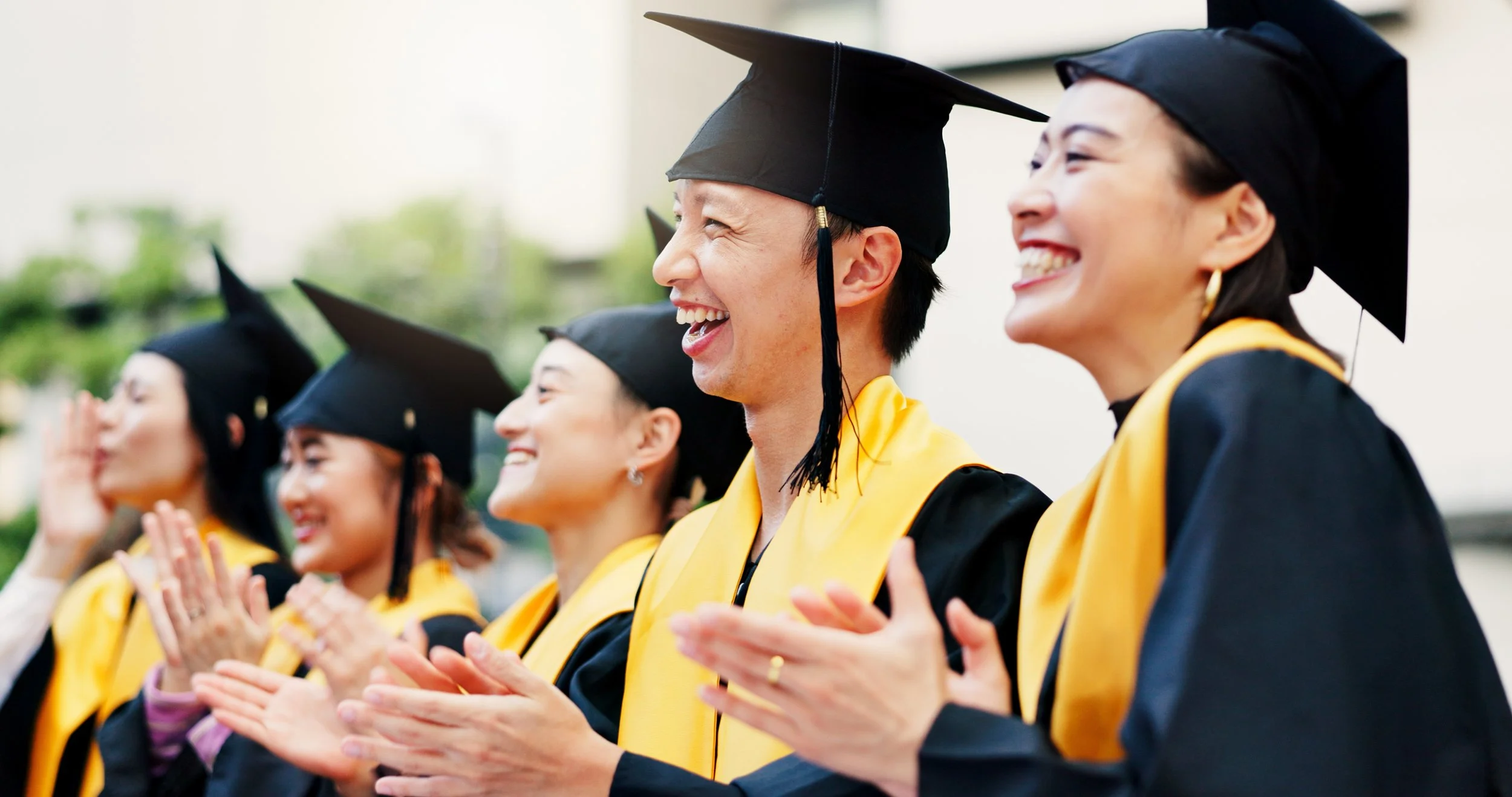 Group of graduates wearing black caps and gowns with yellow stoles, smiling and clapping at a graduation ceremony.