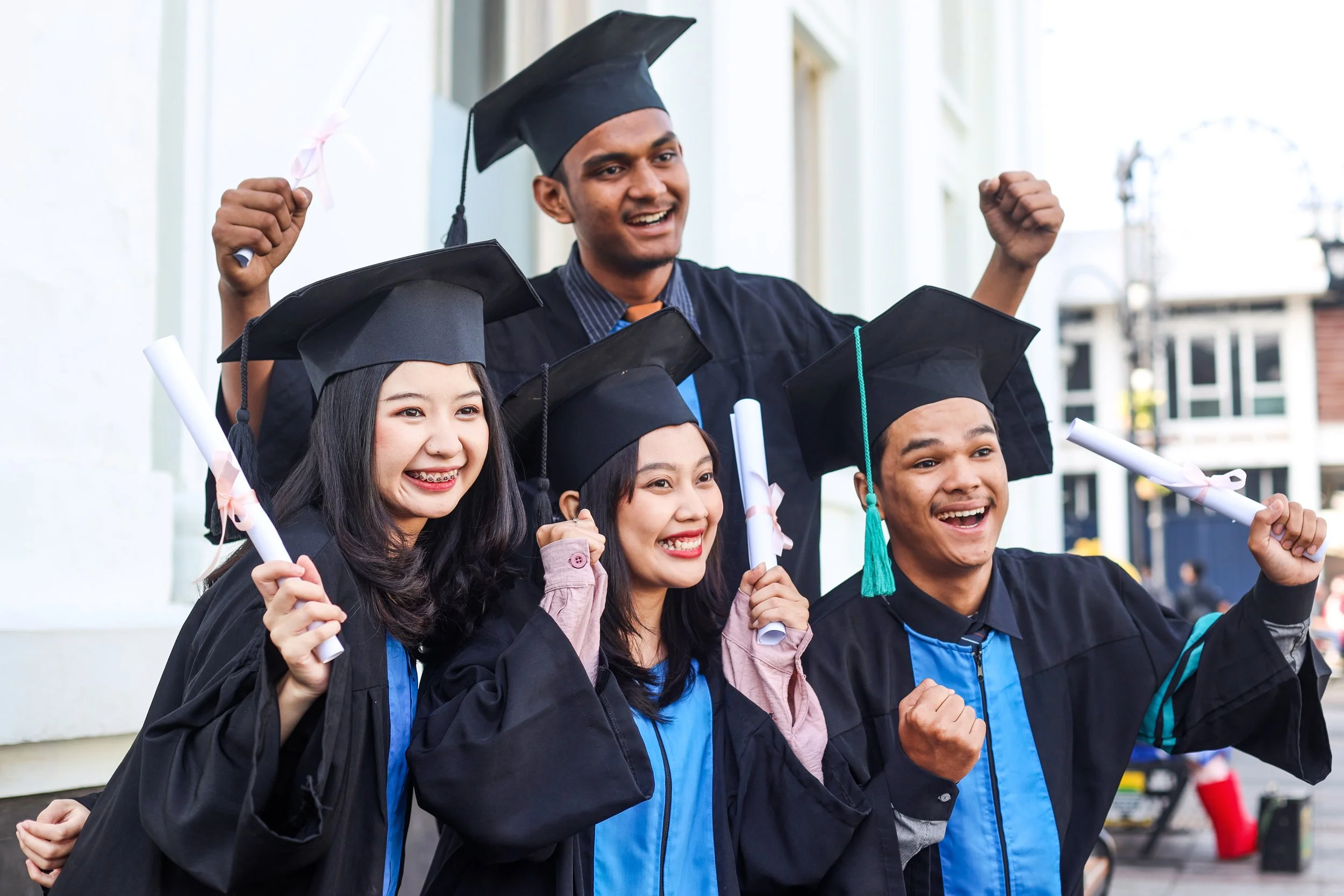 Group of four young graduates in caps and gowns celebrating outside, holding diplomas, with smiling faces and raised fists.