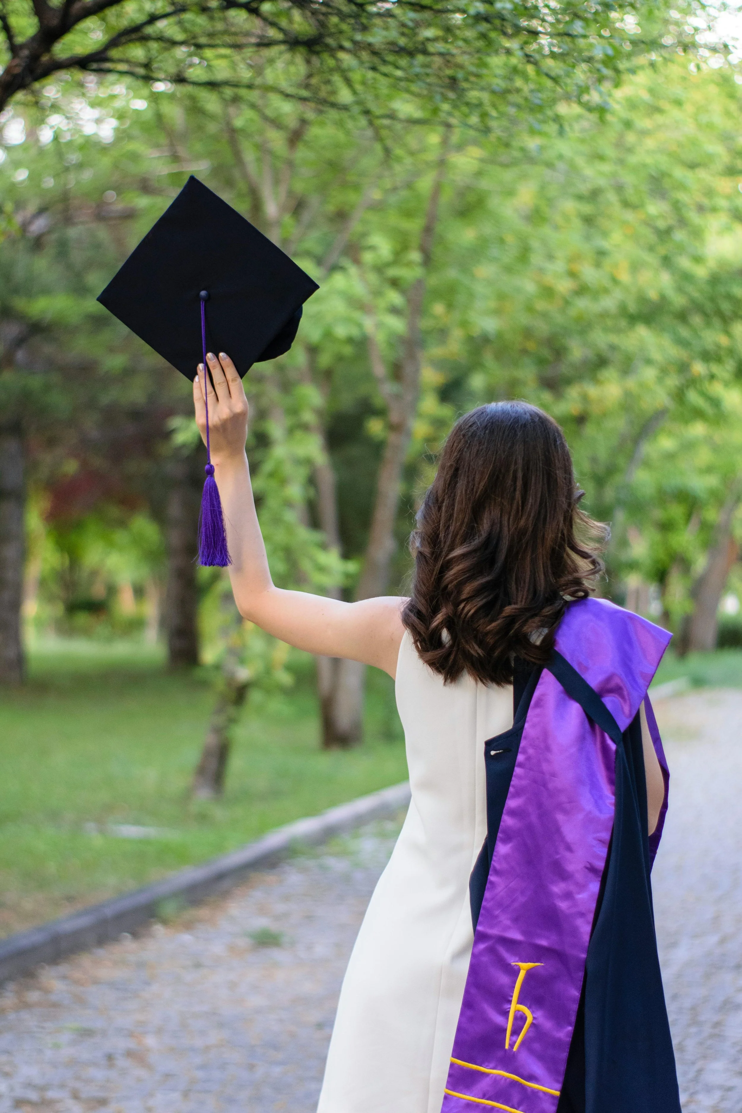 A young woman in a white dress celebrating her graduation outdoors, holding her graduation cap in the air with her right hand, and wearing a purple and navy graduation stole over her shoulder. The background features lush green trees and a pathway.