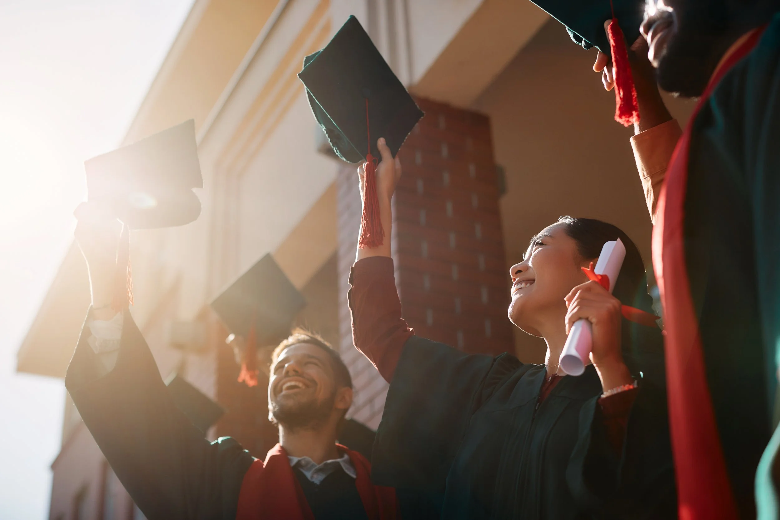 Three graduates in caps and gowns celebrating outdoors, smiling and throwing their caps into the air at sunset.