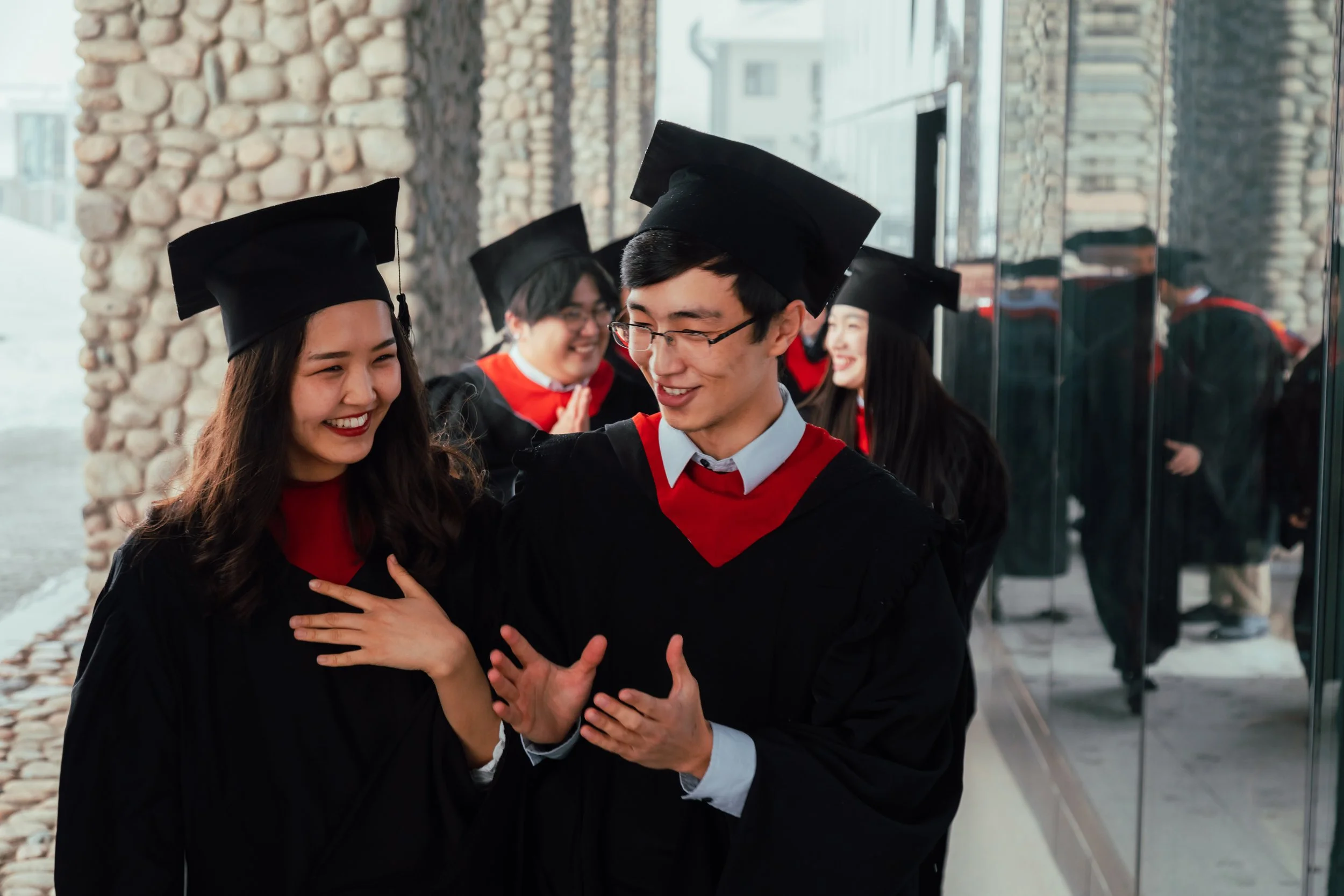 Group of graduates in black caps and gowns smiling, standing outside near a glass wall, celebrating their graduation.